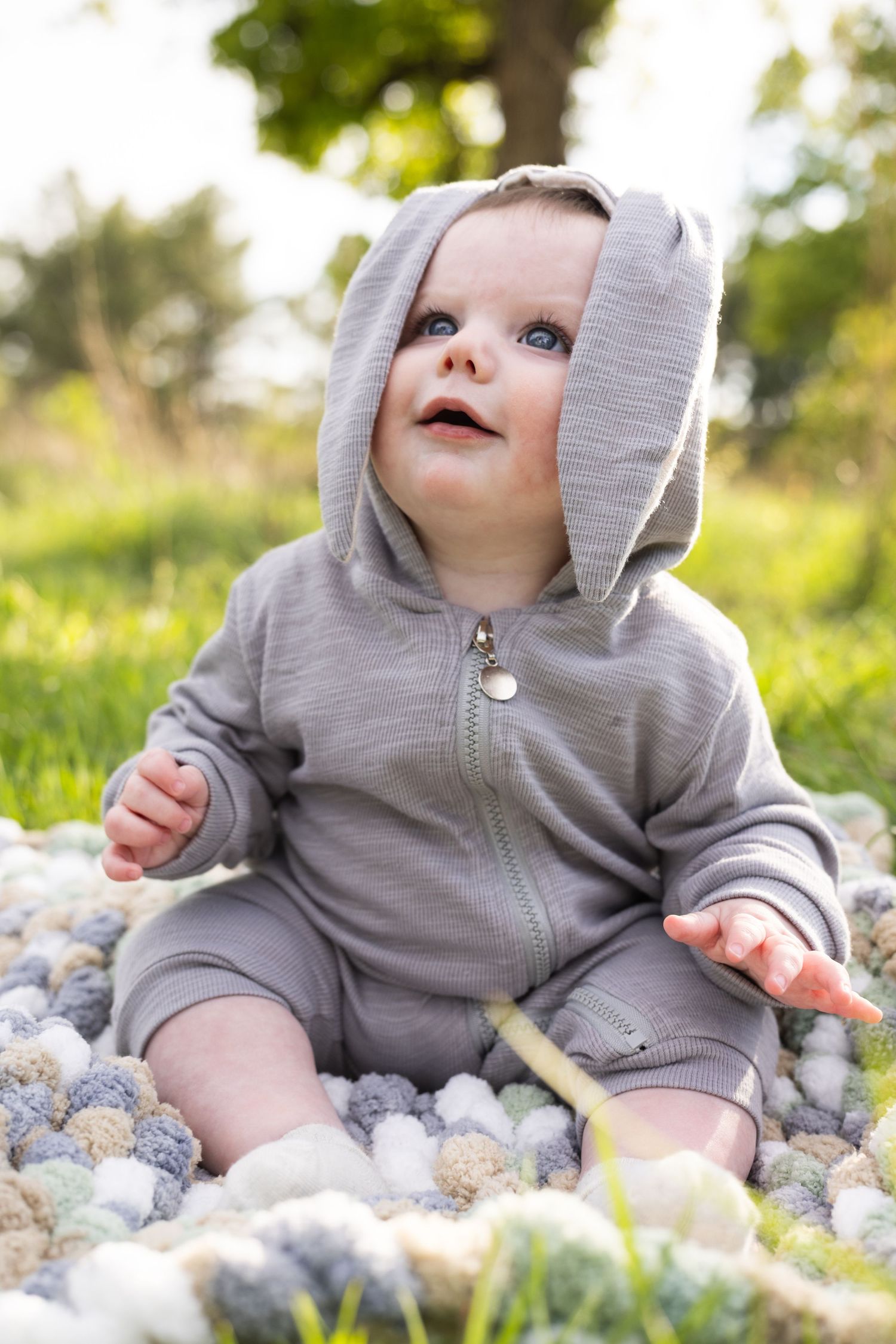 Adorable baby in gray bunny hoodie sitting on colorful felt balls in a sunny green garden.
