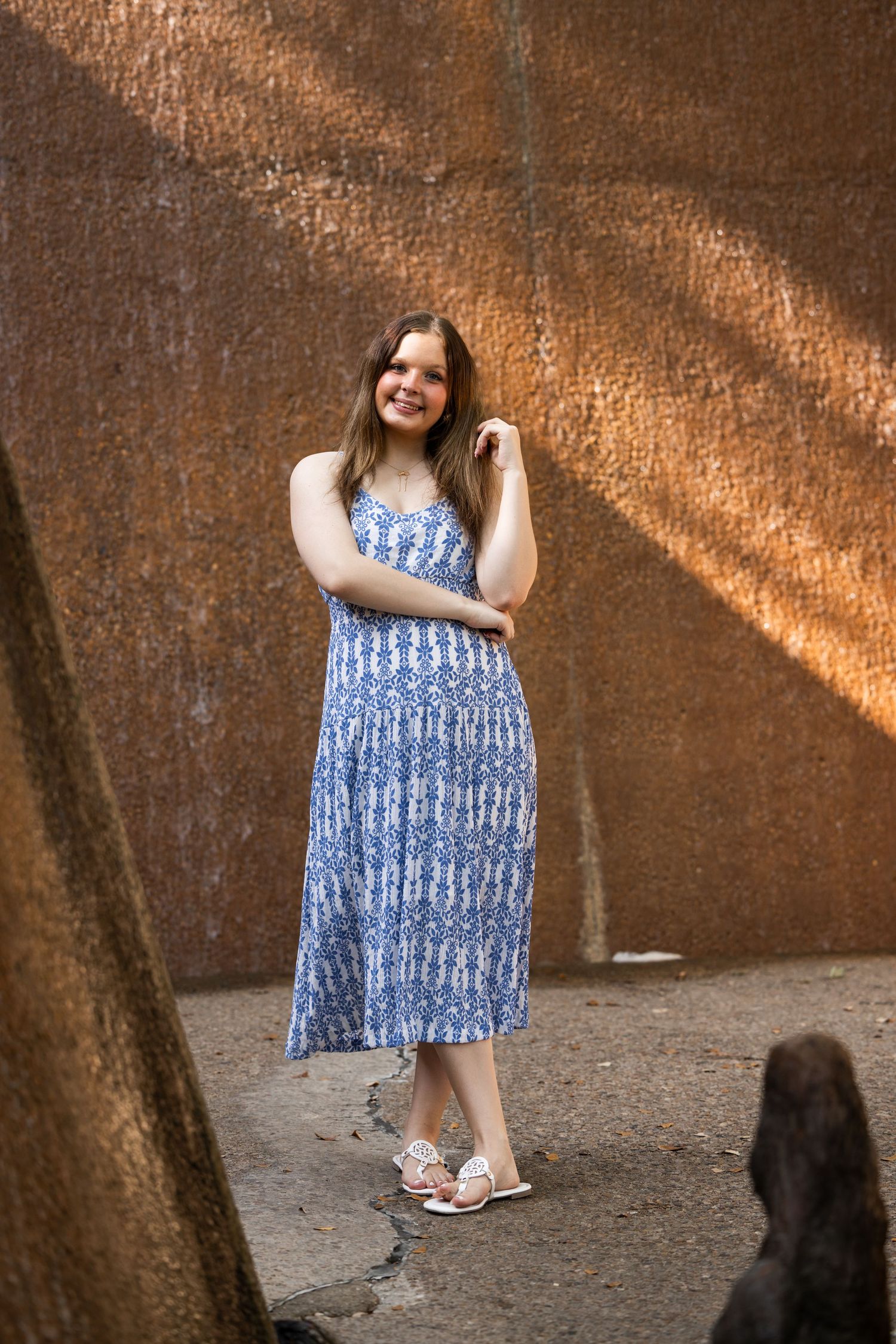 Young woman in blue floral midi dress and sandals smiling against rustic metal wall backdrop.