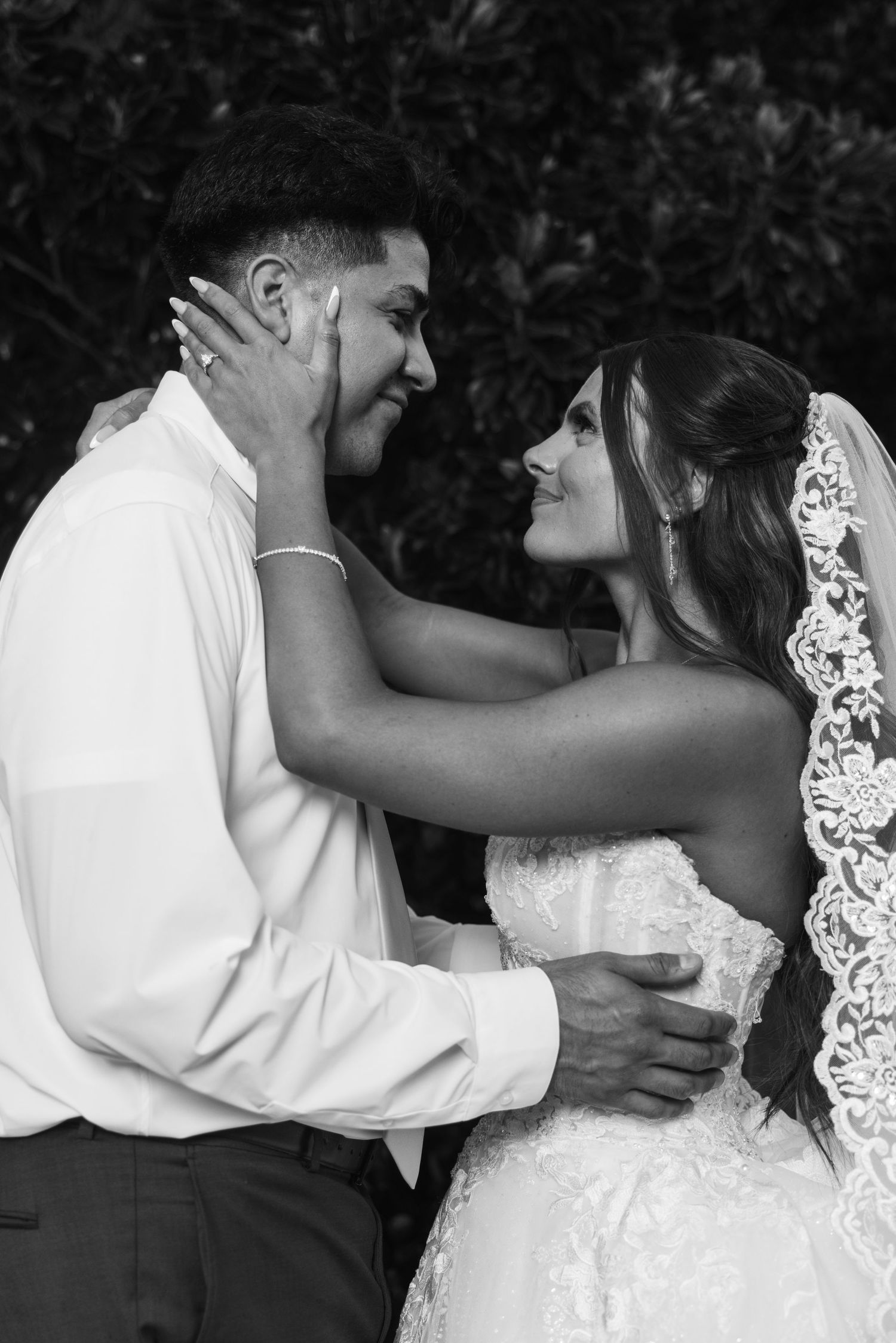 Black and white wedding photo of bride and groom gazing at each other, bride in lace gown and veil, groom in white shirt.