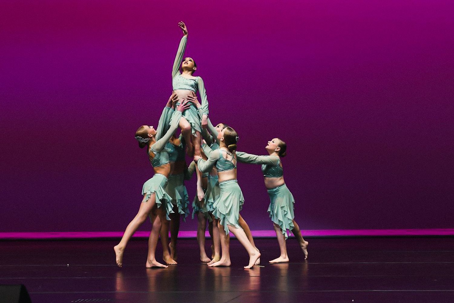 Dancers in teal and white costumes perform a dynamic contemporary dance routine on a dark stage with purple lighting.