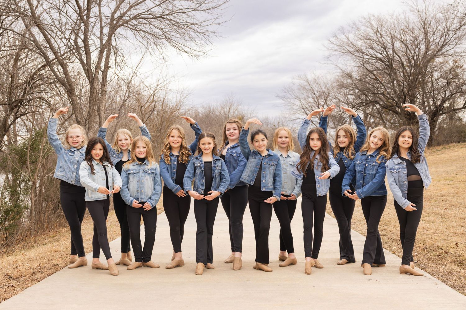 Group of women wearing matching denim jackets and black pants pose together on a path during autumn.