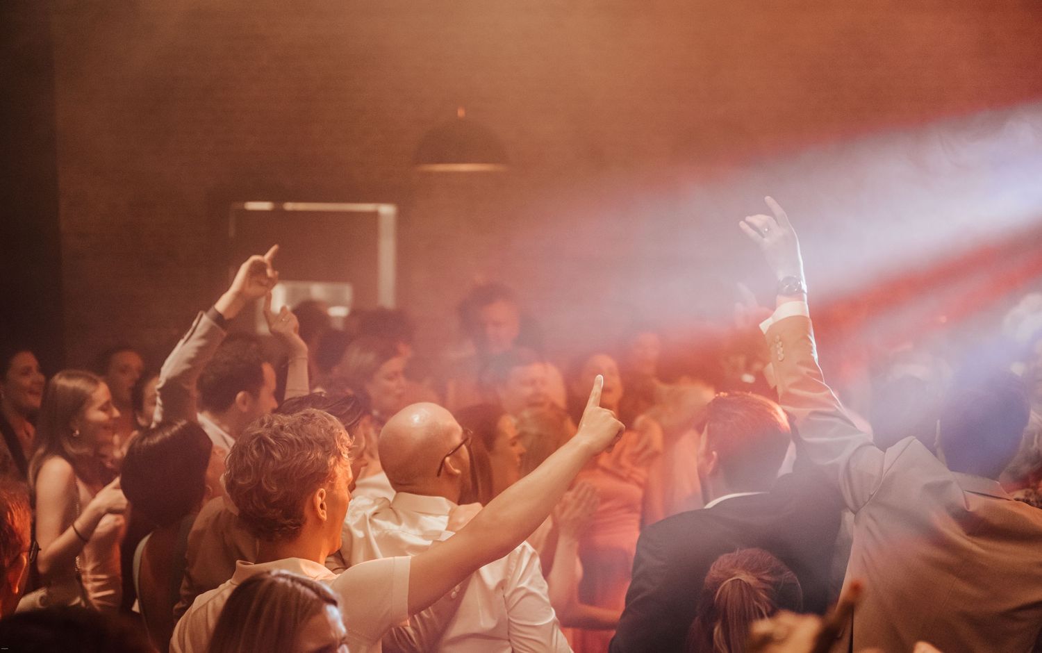 Crowd of wedding guests dancing and celebrating on a dimly lit dance floor with red lighting effects.