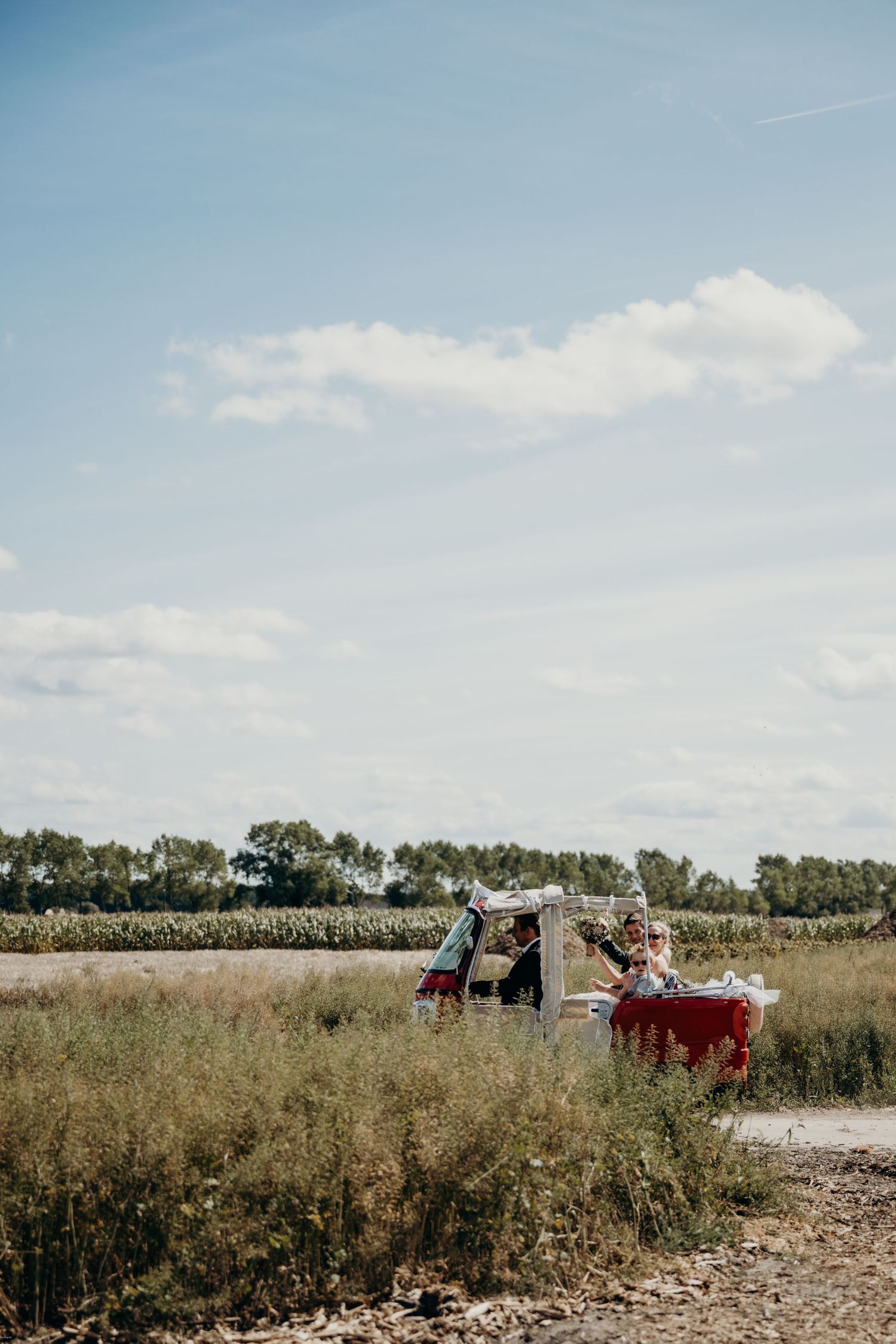 Wedding party riding in a vintage vehicle through a rural field on a sunny day.