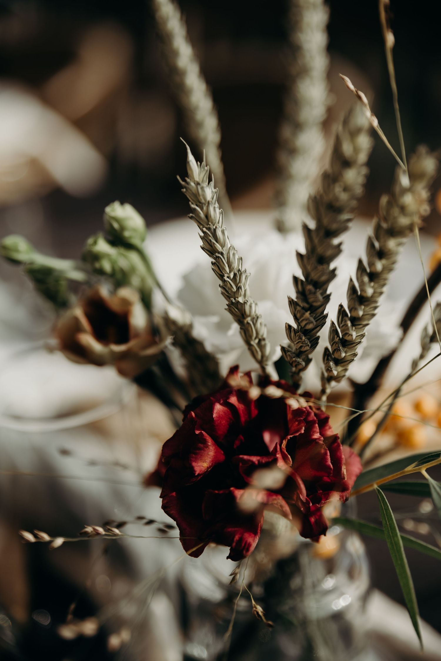 Deep red flowers and dried stems create a moody floral arrangement in dark lighting.