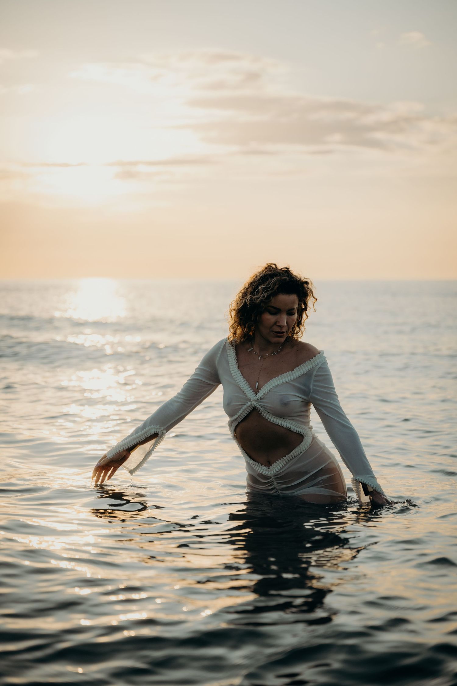 Figure in white clothing stands in ocean water during golden hour sunset with waves gently lapping.