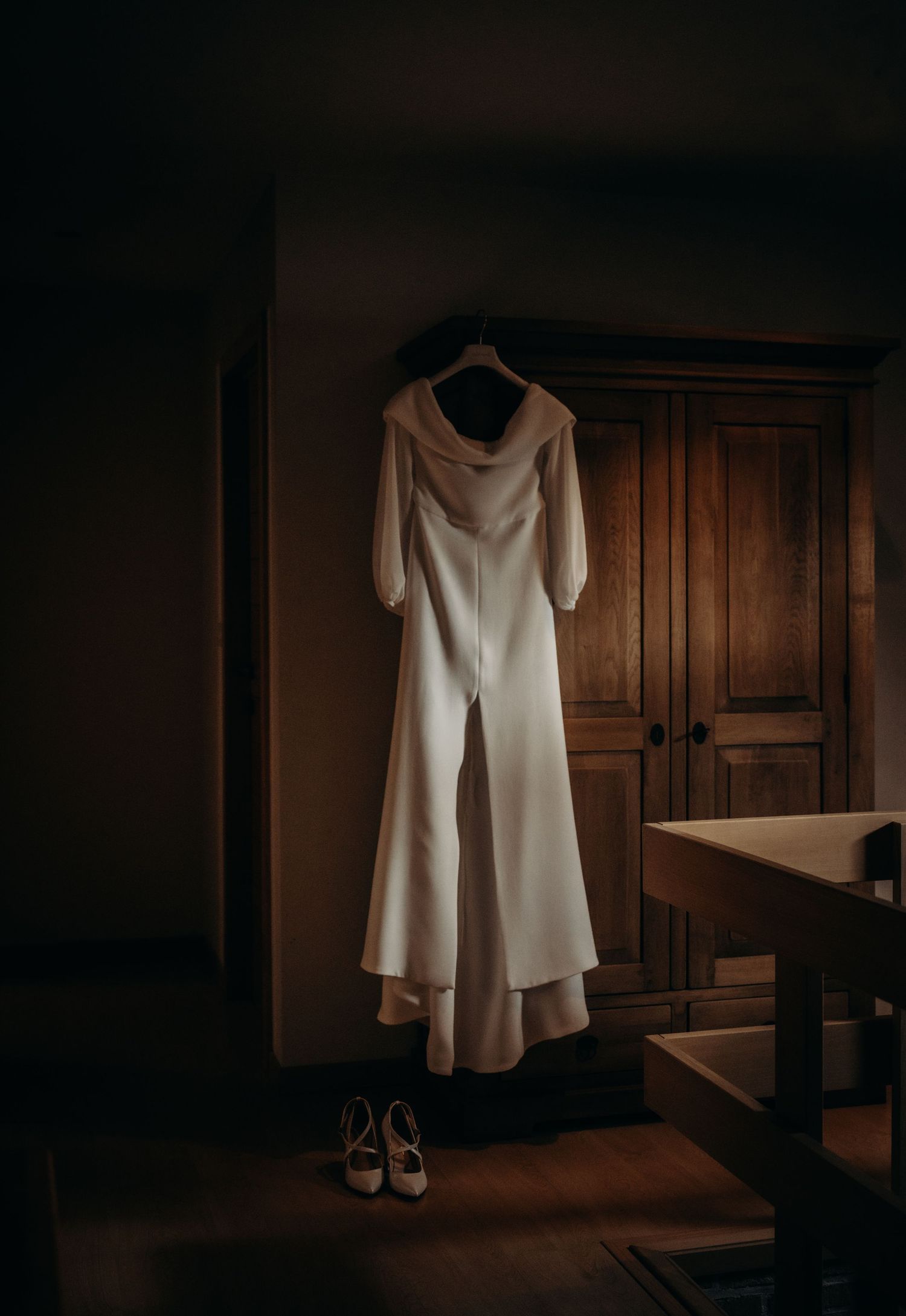 Elegant white wedding dress hanging in a dark wooden room with dramatic lighting.