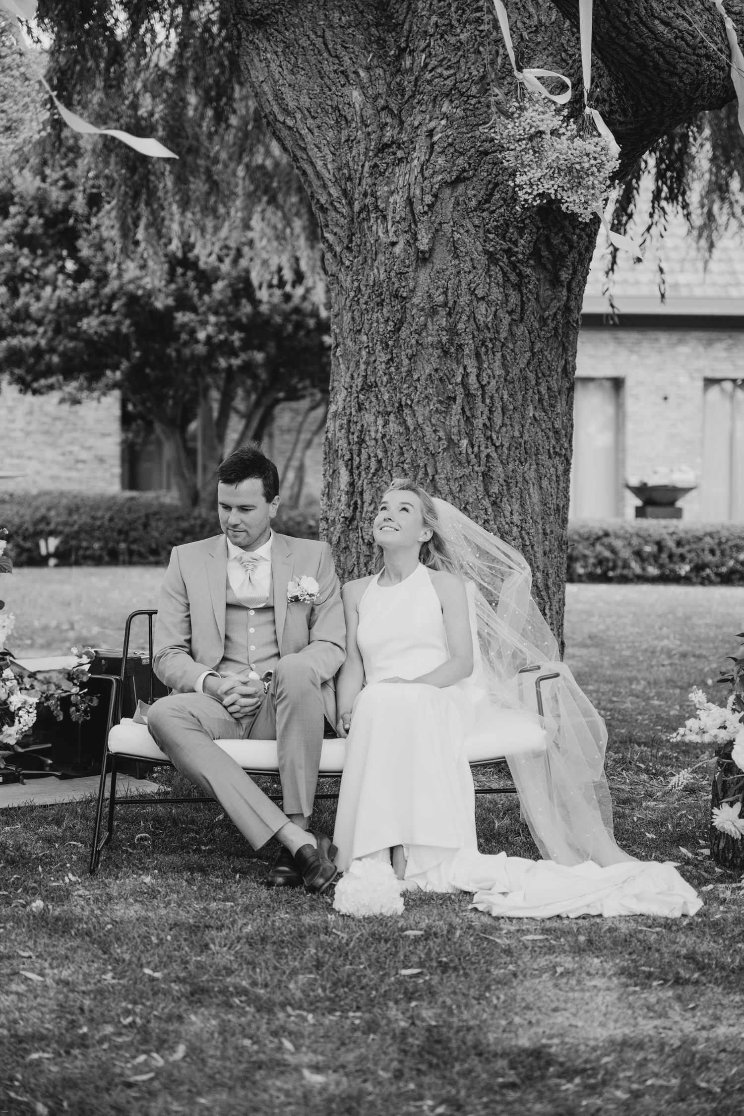 A couple sits together under a large tree during their outdoor wedding ceremony.