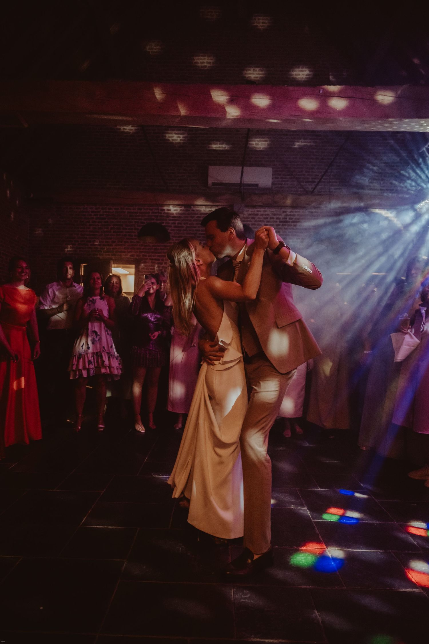 Wedding guests dancing in formal attire under colorful disco lights in a dark reception venue.