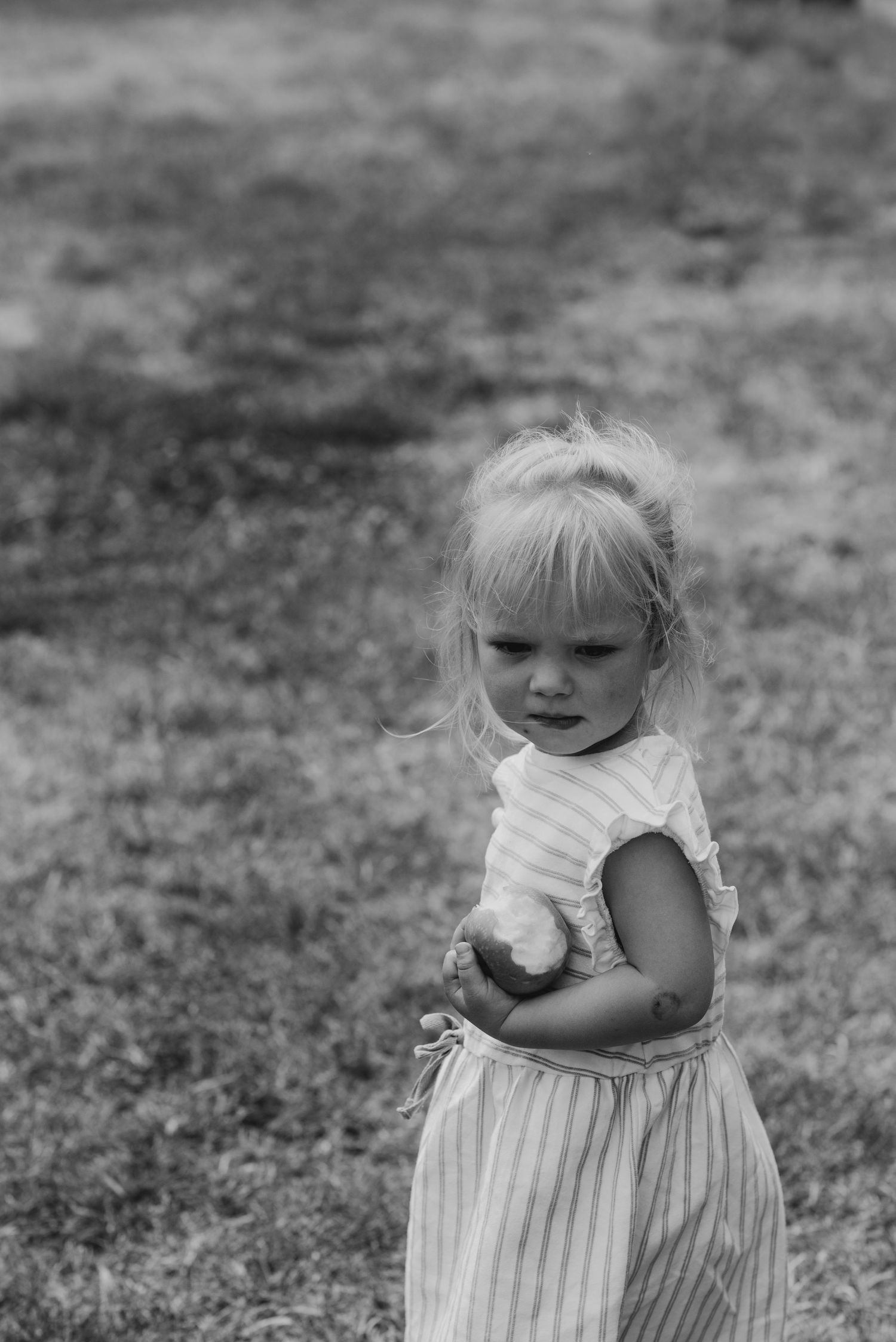 A young child in a striped dress stands thoughtfully in a grassy area, captured in black and white photography.