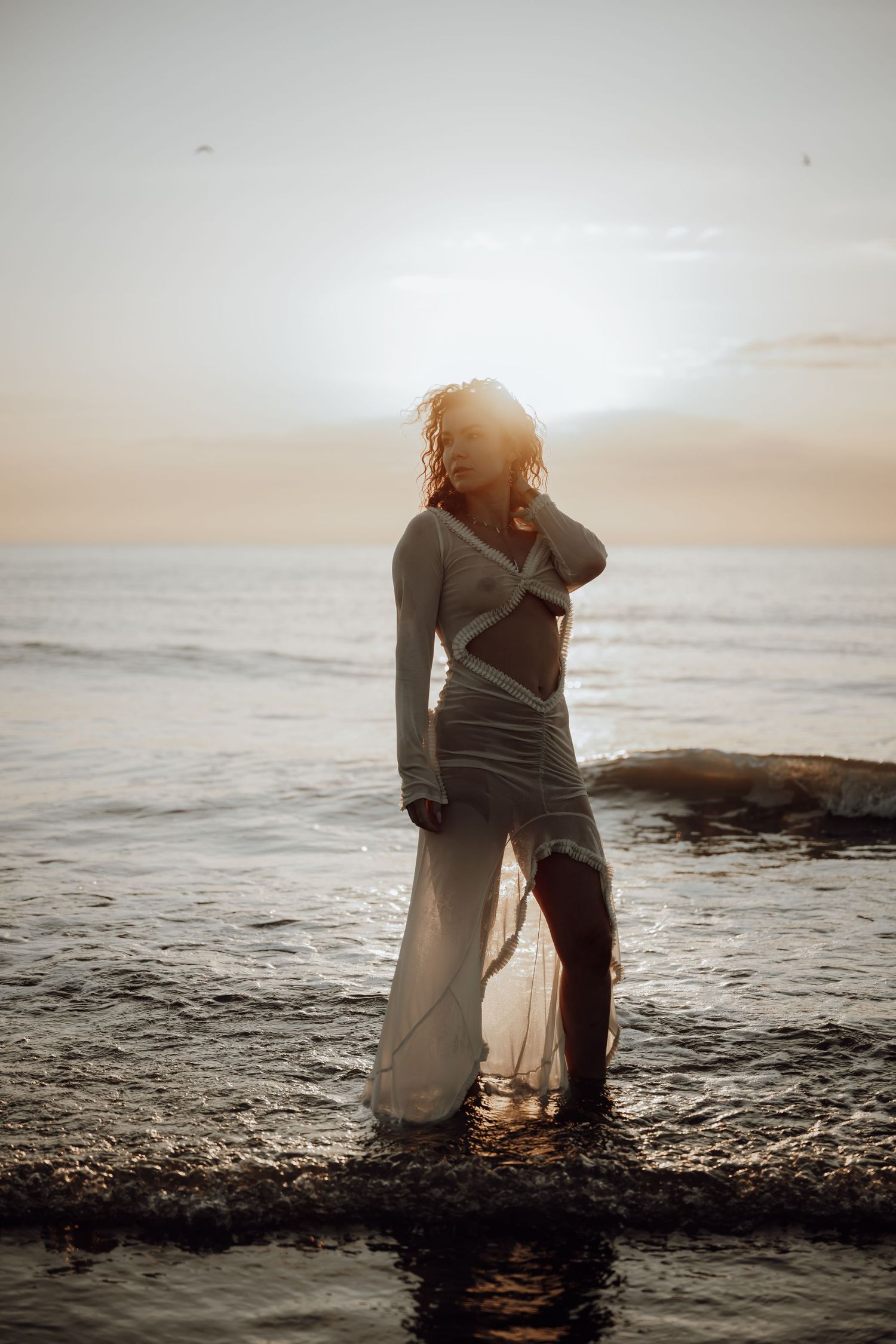 Person in flowing white dress crouches at the ocean's edge during golden hour sunset.