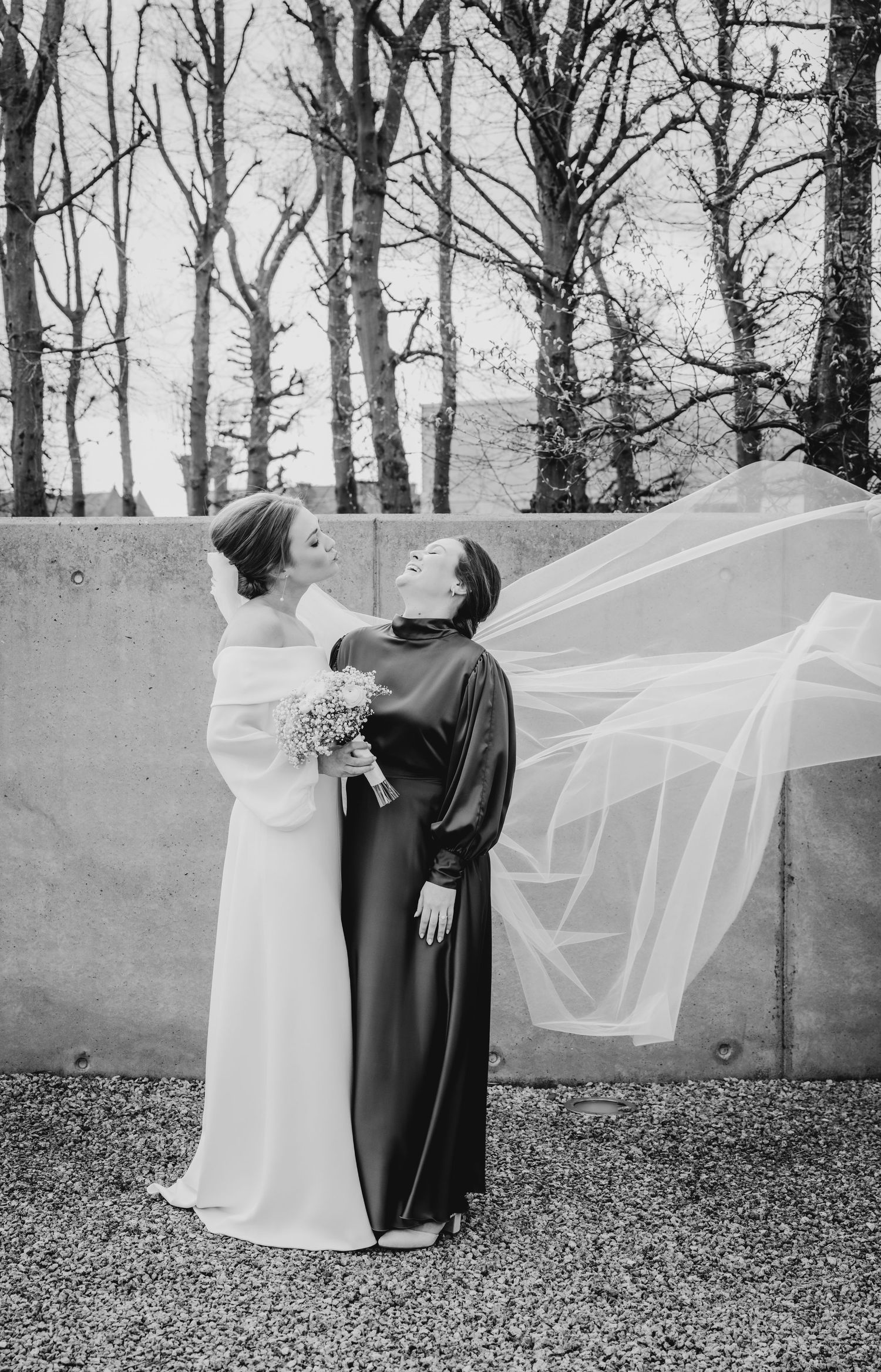 Winter wedding photos with flowing veil against bare trees and stone wall.