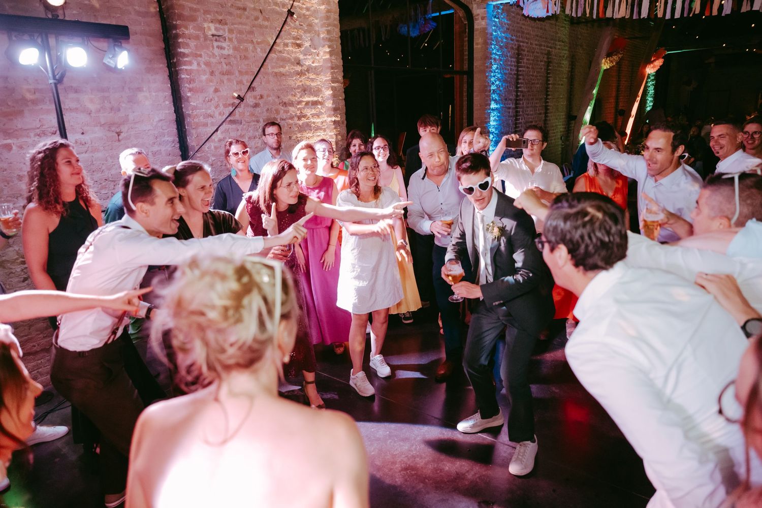 Wedding guests dance and celebrate on a crowded dance floor with exposed brick walls and festive lighting.