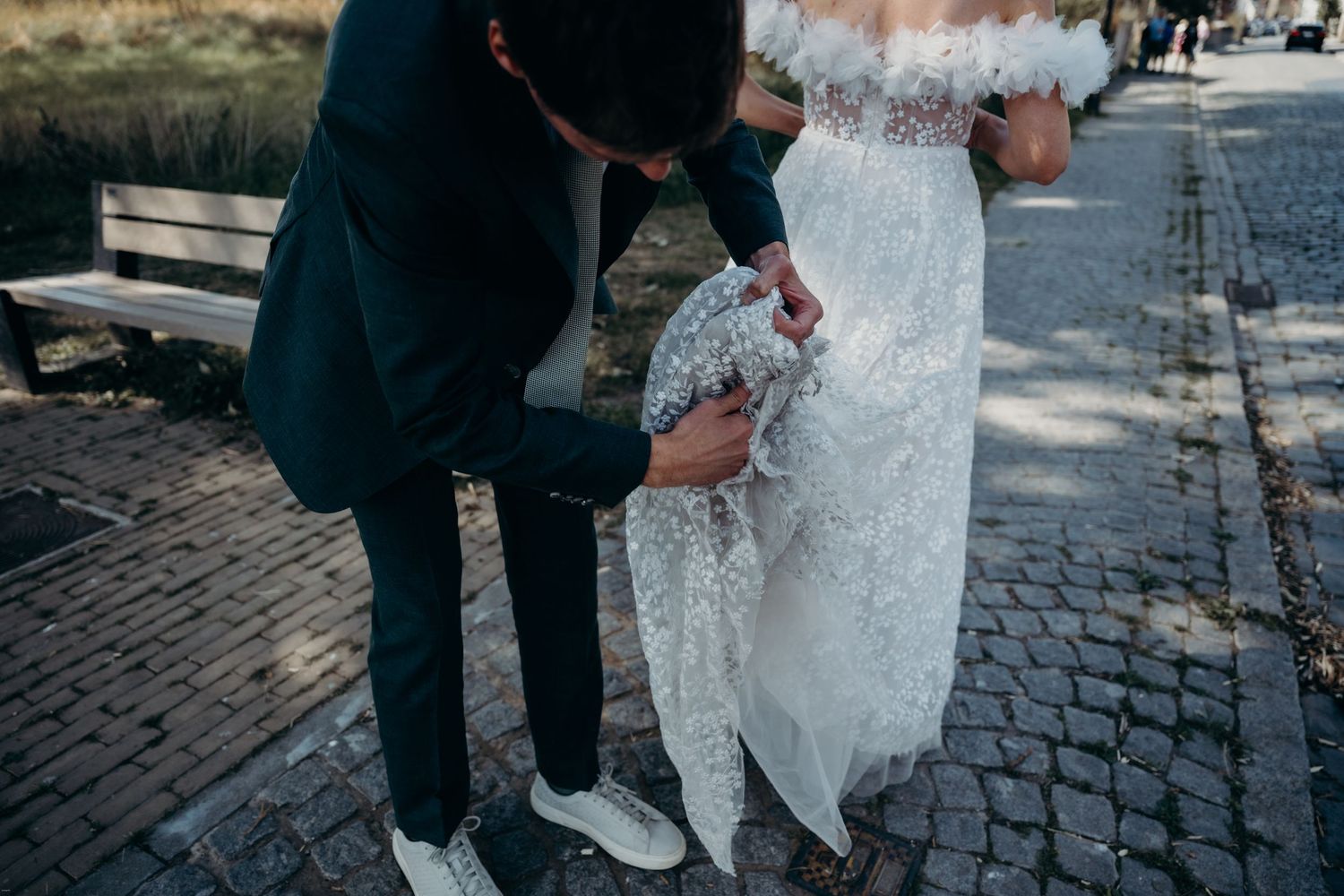 Person in formal wear helping adjust a white lace wedding dress on cobblestone street.