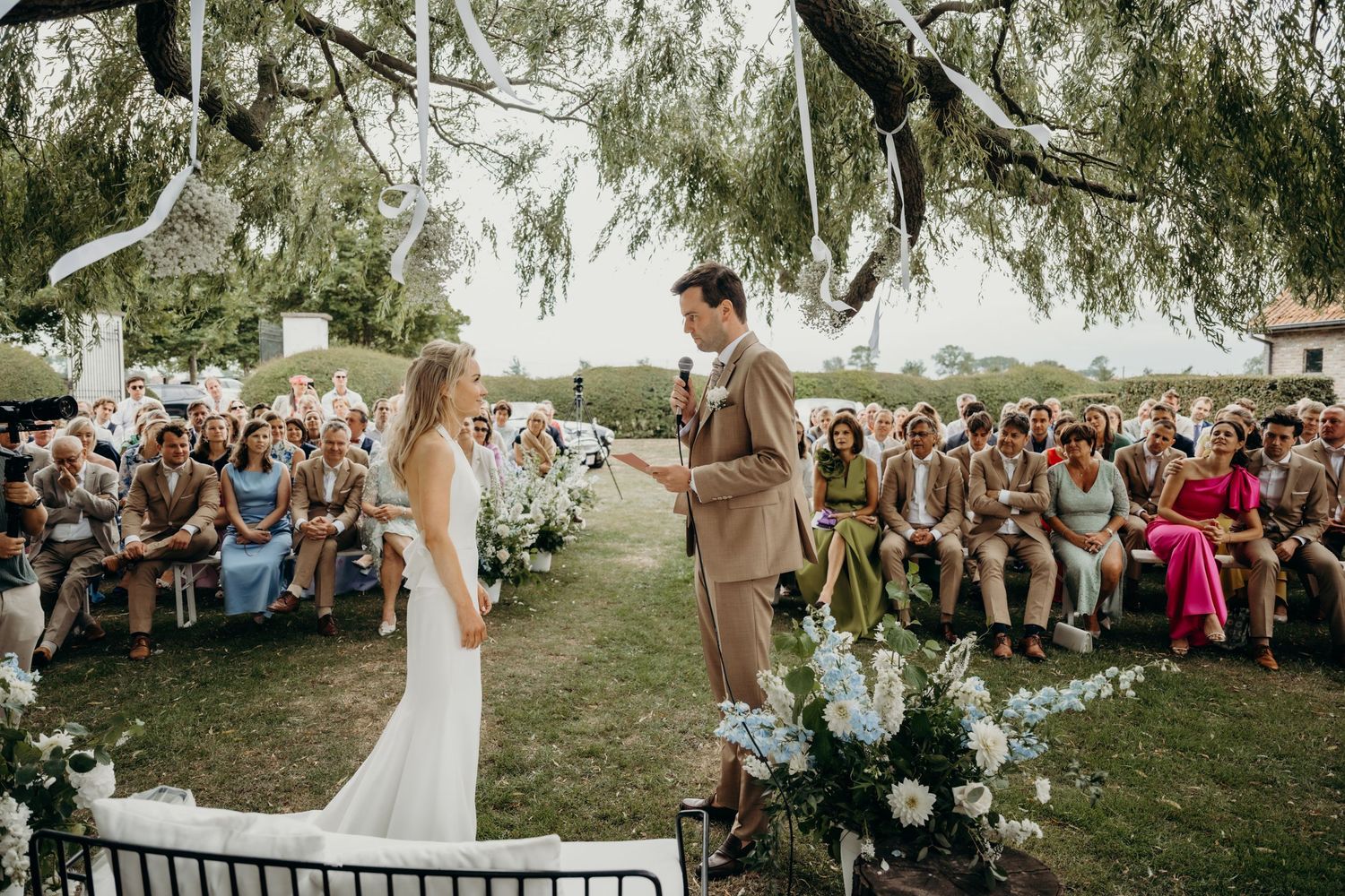 An outdoor wedding ceremony with guests seated on wooden chairs under a willow tree.