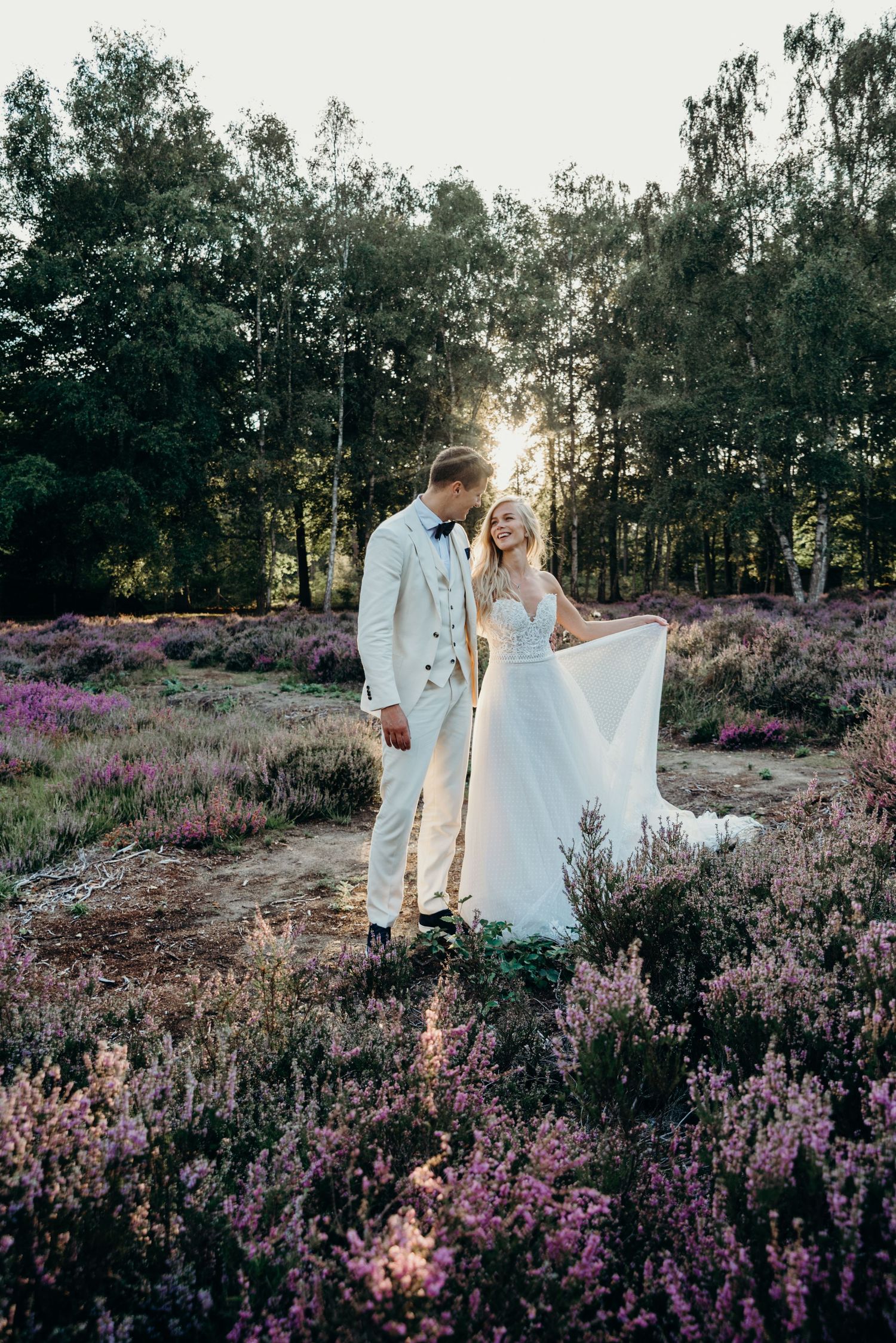 Wedding couple standing in field of purple heather with sunlit forest backdrop.