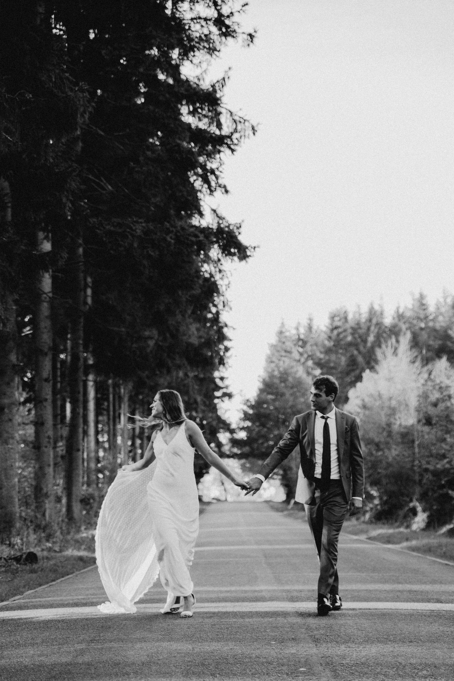 Black and white photo of couple walking down tree-lined path with flowing wedding dress.