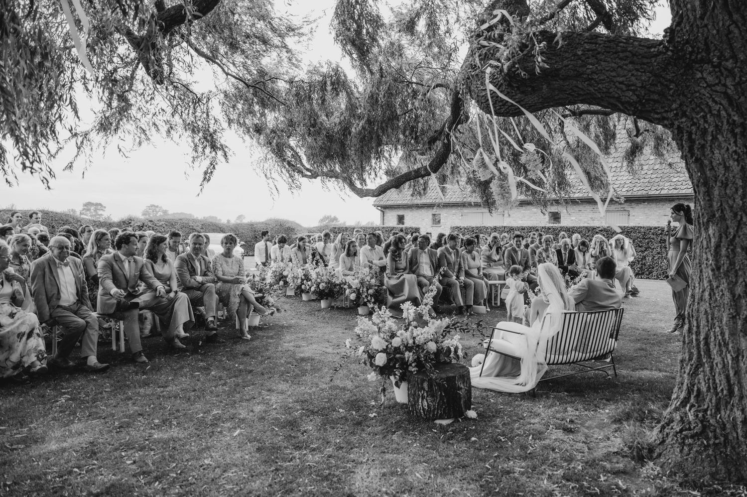 Black and white photo of an outdoor wedding ceremony with guests seated under a large tree.