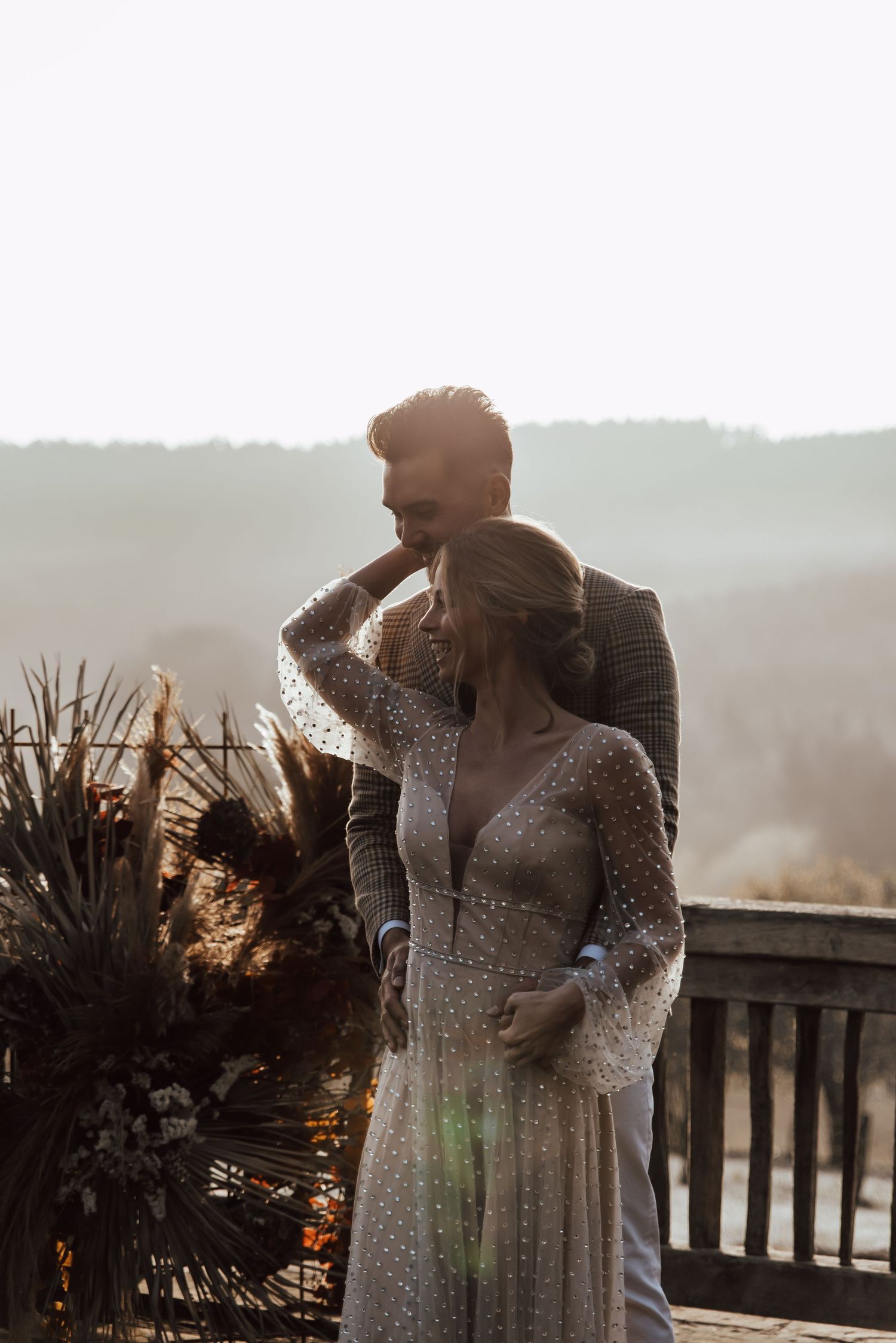 Couple in wedding attire share intimate moment on deck overlooking misty mountain landscape.