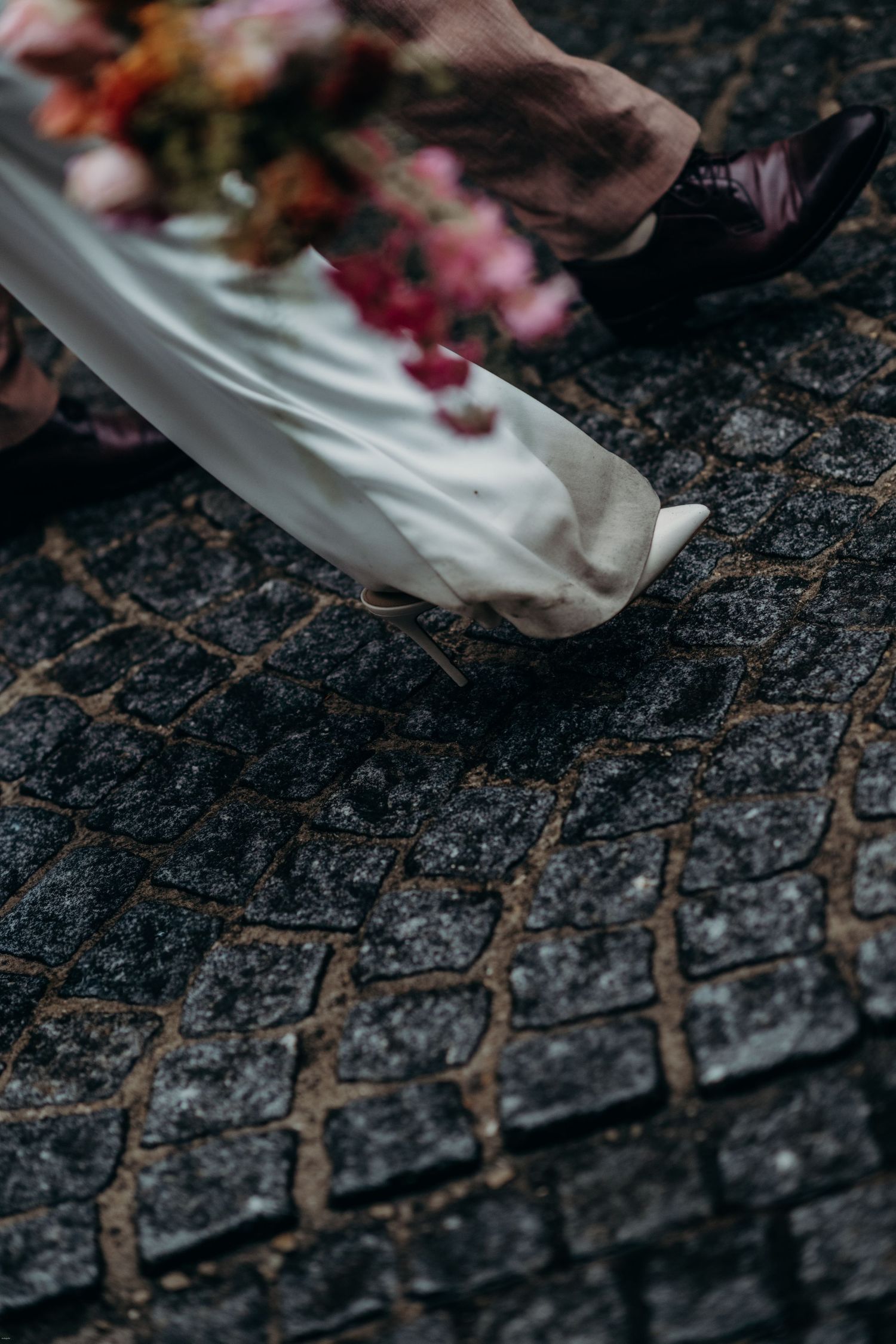 Wedding dress hem trailing over dark textured cobblestones with flower petals.