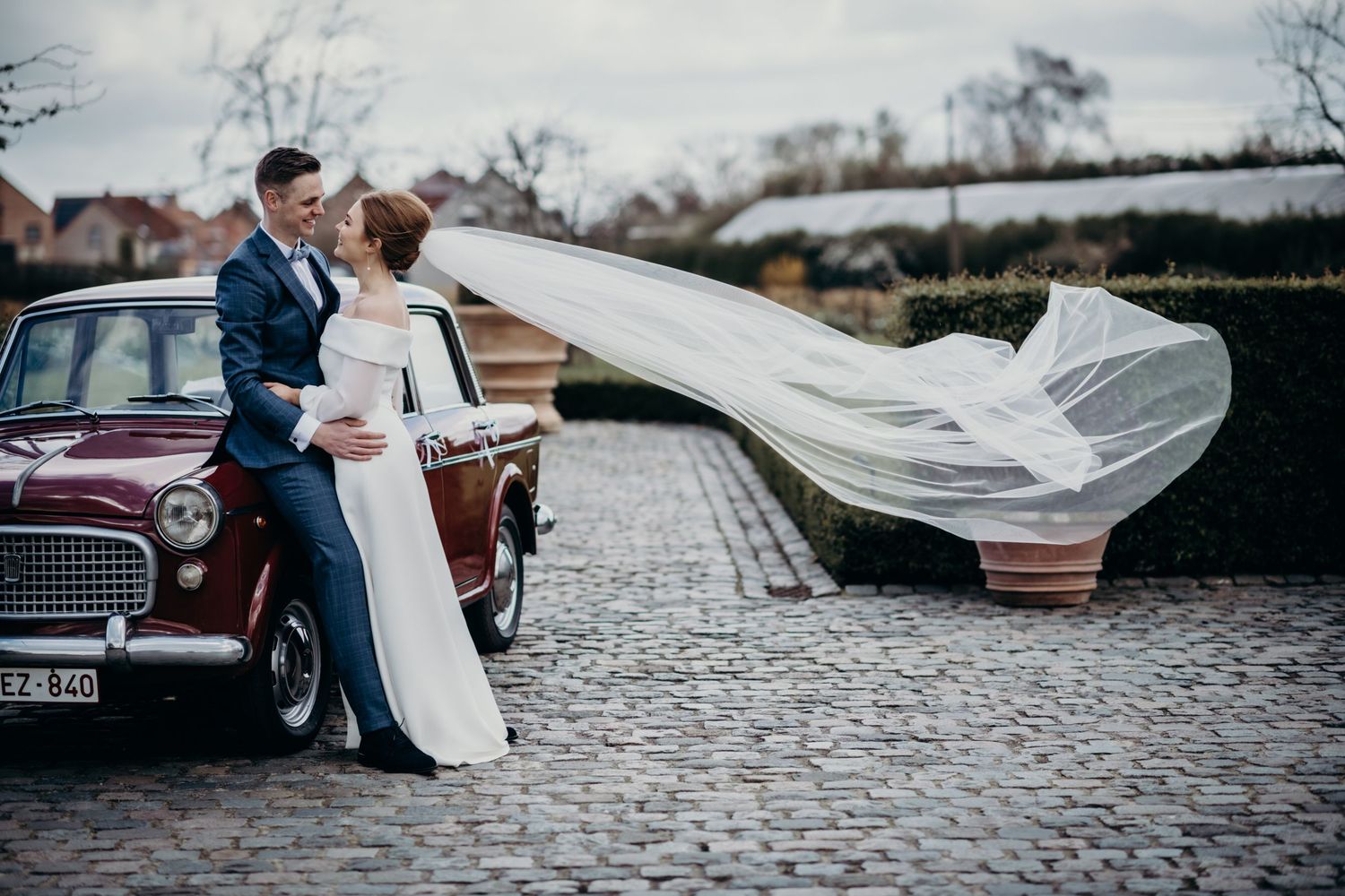Wedding couple posing with flowing veil on cobblestone street with vintage car.