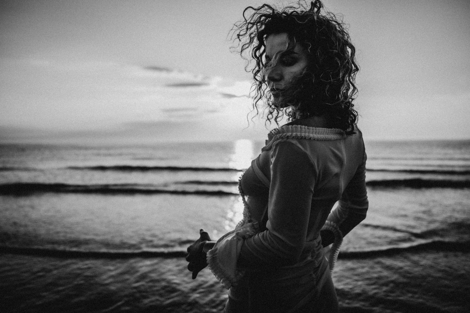 A silhouetted figure stands against a dramatic ocean sunset creating a moody black and white beach scene.