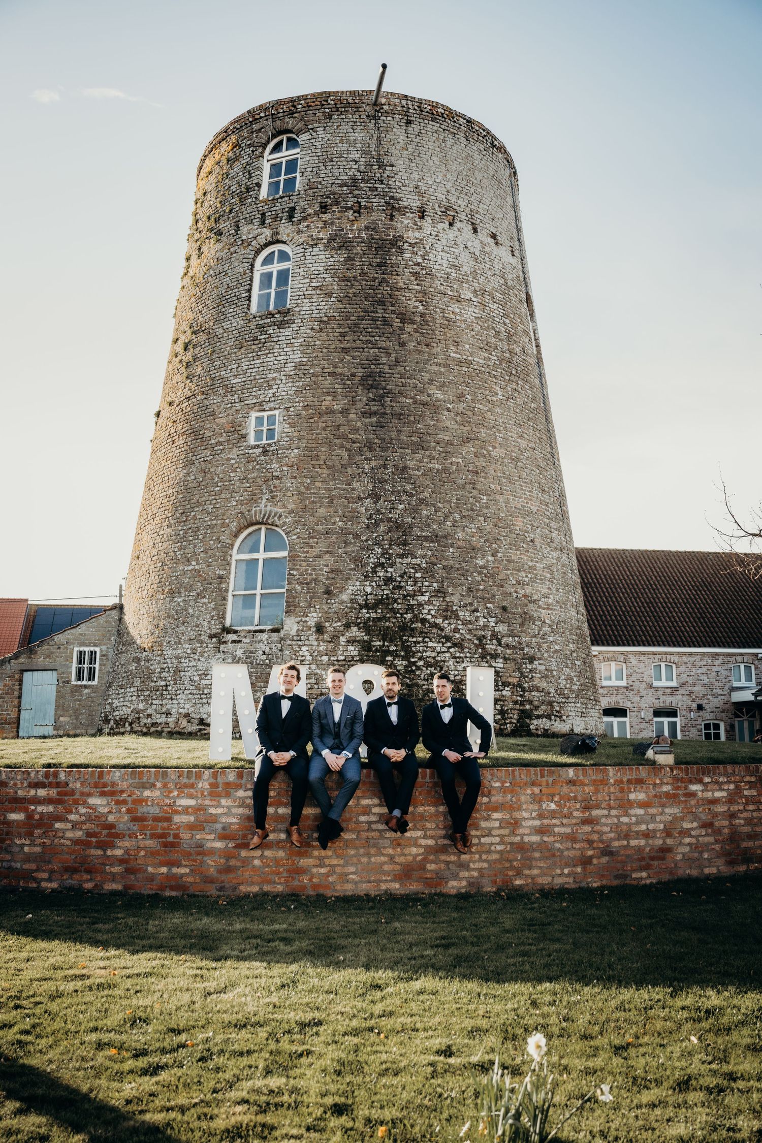 Group photo of wedding party sitting on brick wall in front of stone windmill.