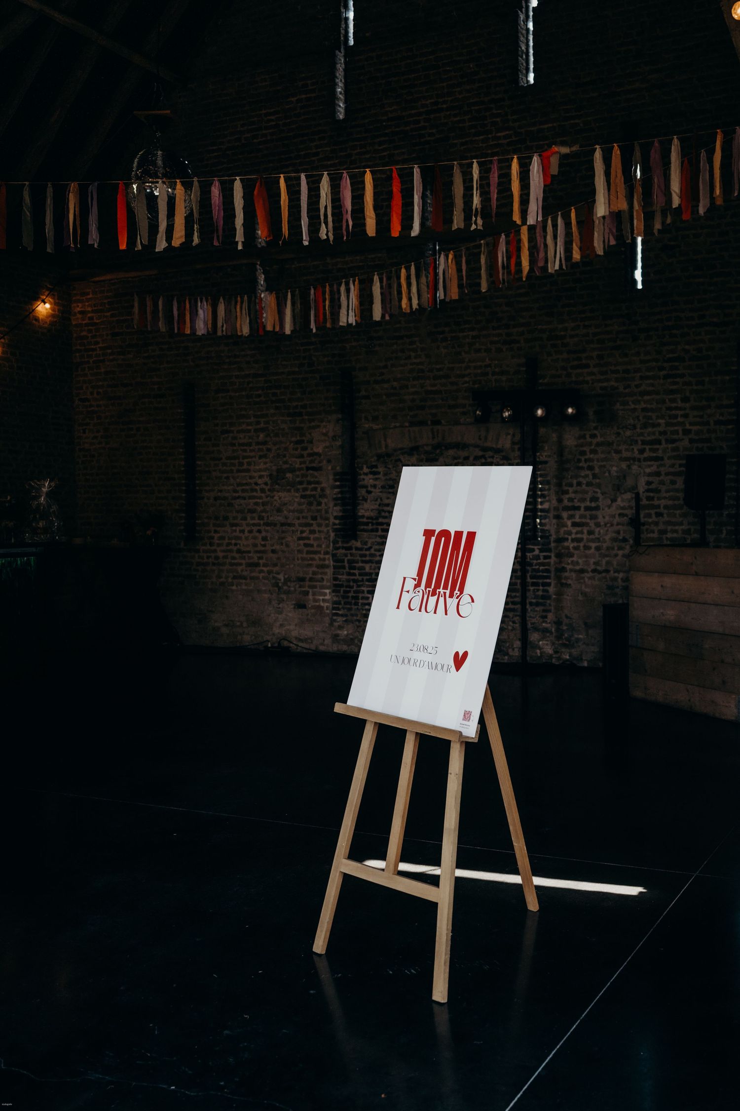 Wedding signage displayed on an easel under decorative hanging ribbons in a brick venue.