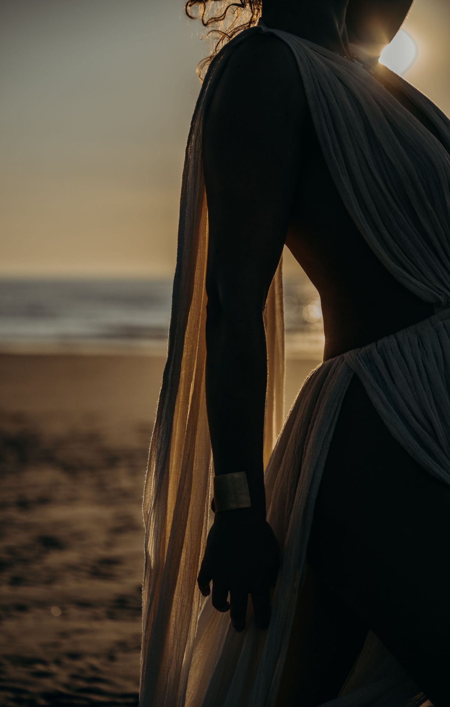 Silhouette of pregnant woman in flowing dress standing on beach at golden sunset.