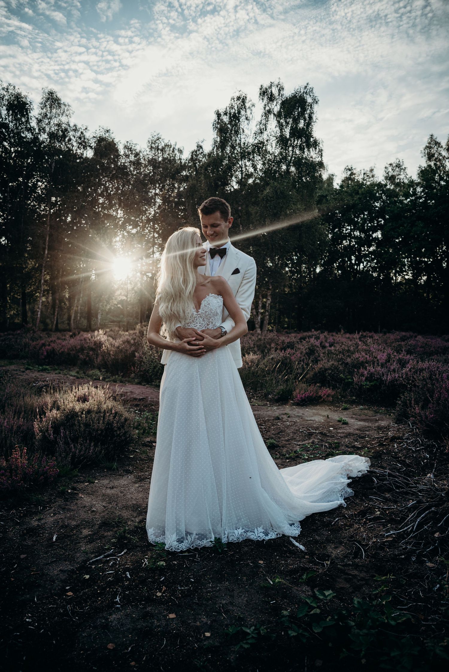 A wedding couple embraces in a dramatic sunset photo with trees and natural landscape.