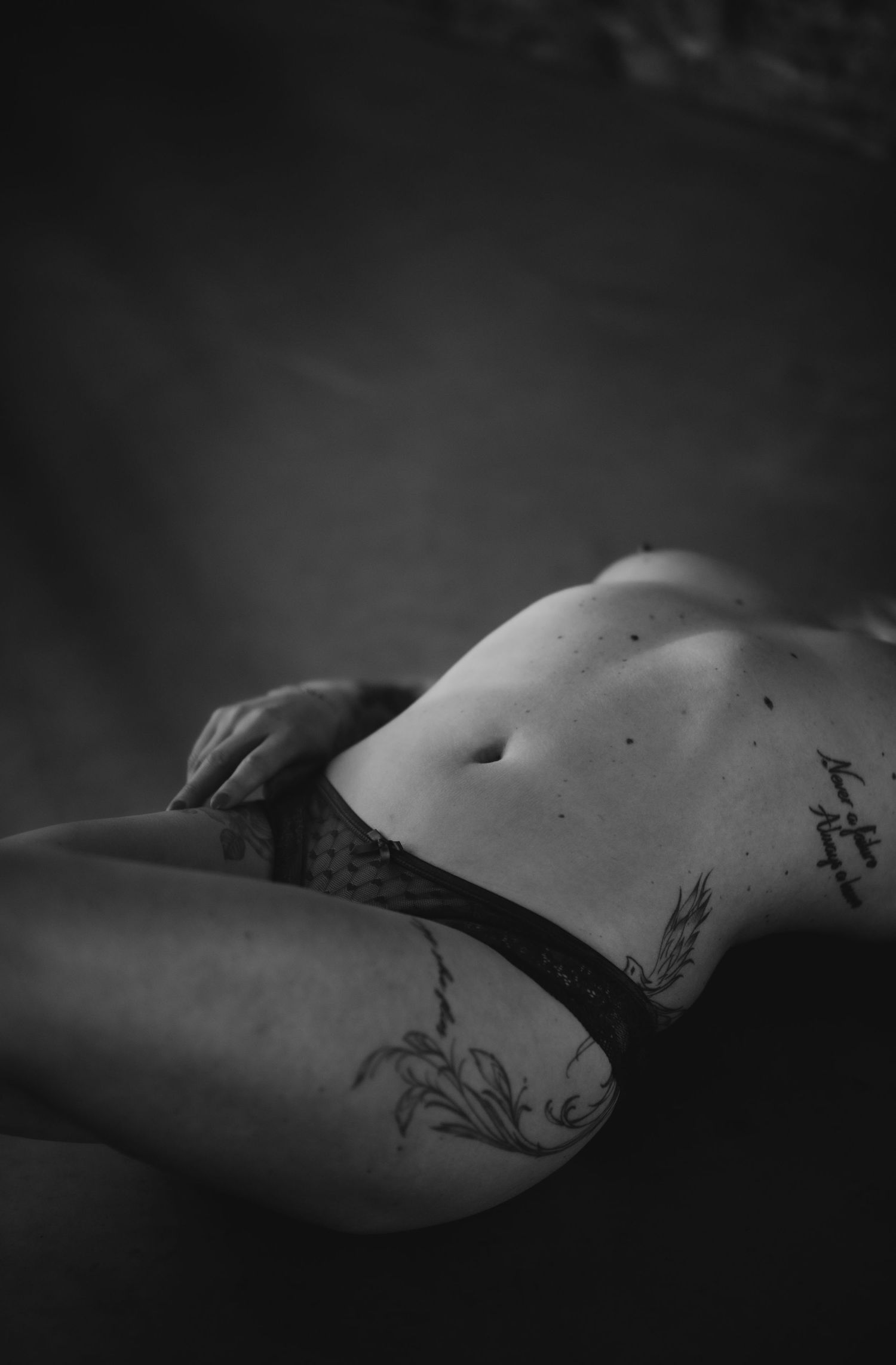 Artistic black and white boudoir photograph showing tattoos on torso and thigh in moody lighting.