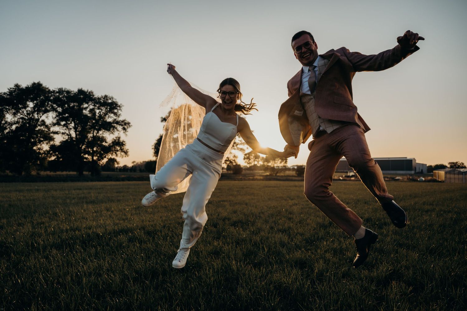 A couple dances and jumps playfully in a golden field at sunset.