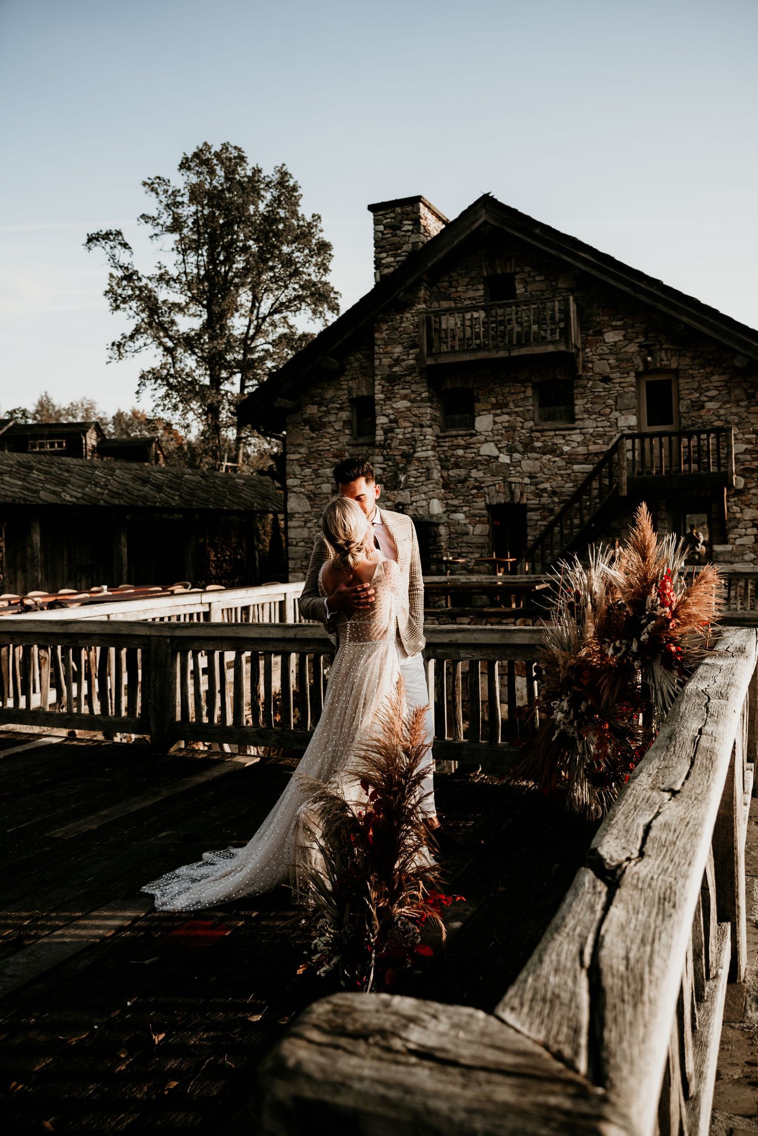 Wedding couple poses at rustic stone venue with wooden deck and autumn floral arrangements.