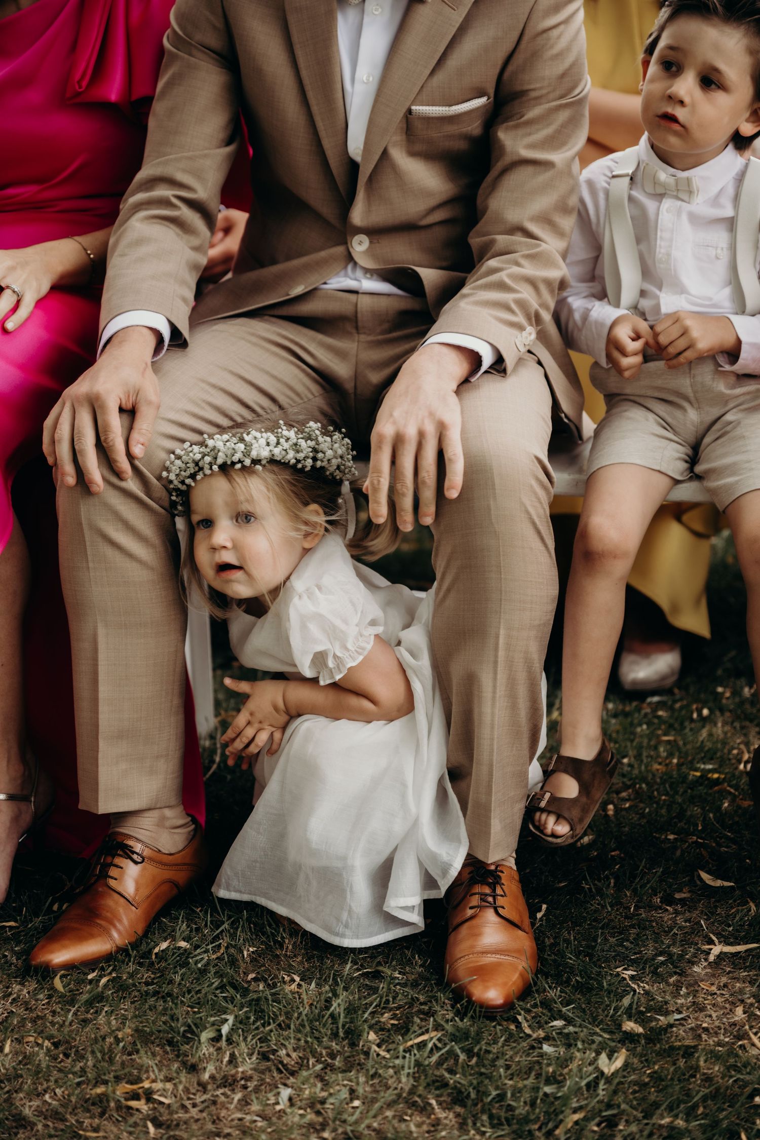 Wedding attendees in formal attire sitting on chairs during an outdoor ceremony.