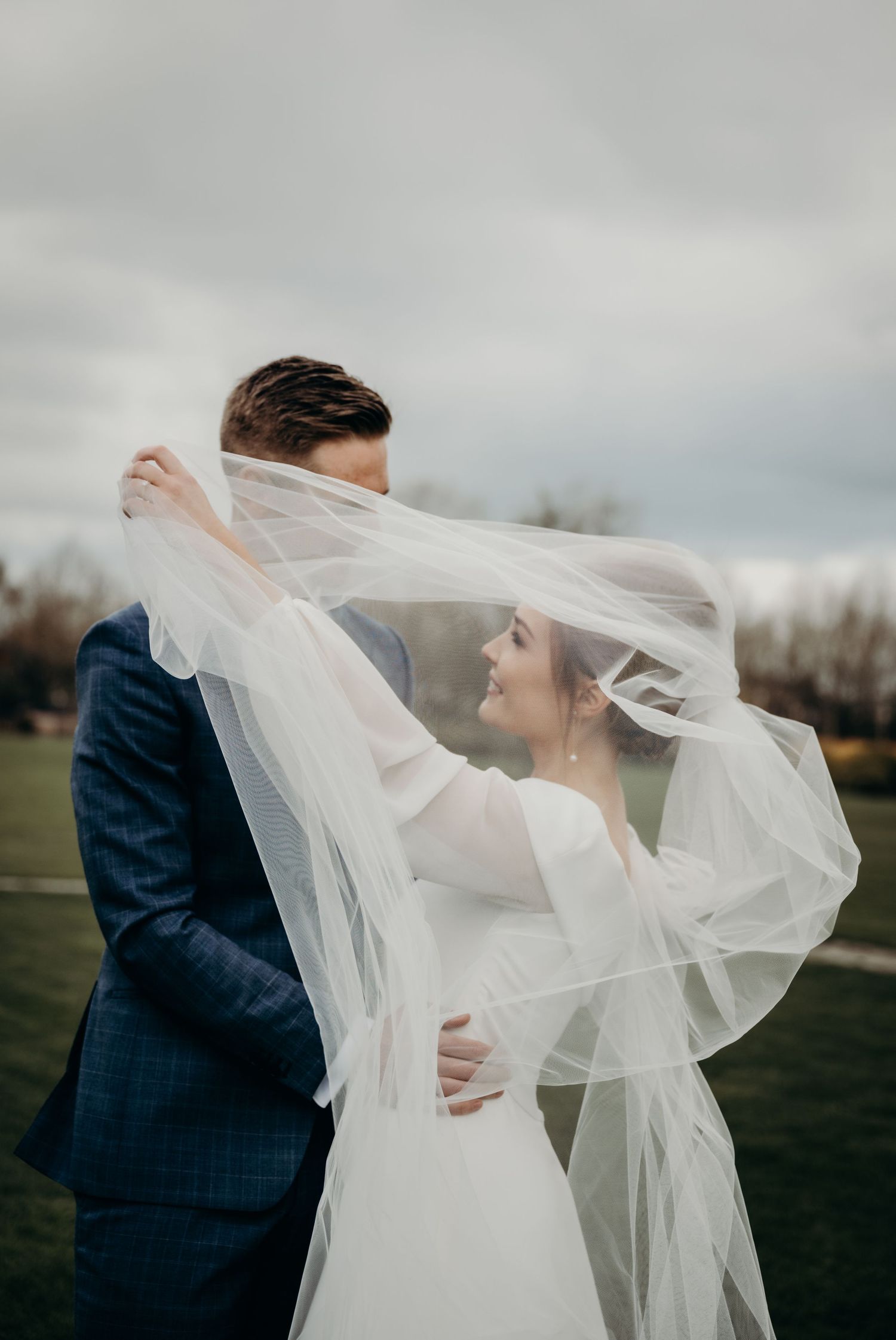 Wedding couple embracing outdoors while veil flows dramatically in the wind.