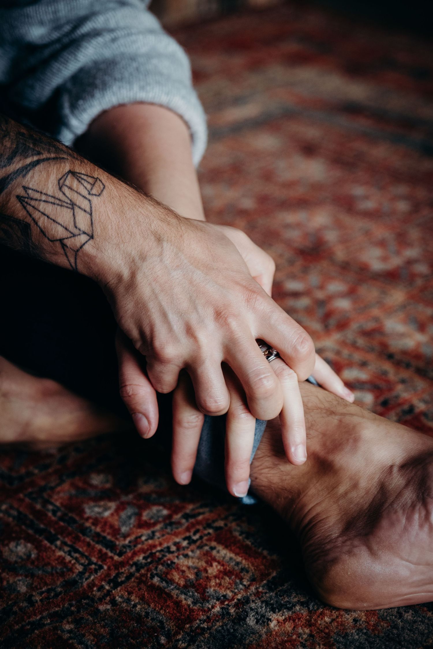 Tattooed hand touching foot on ornate carpet in intimate setting.