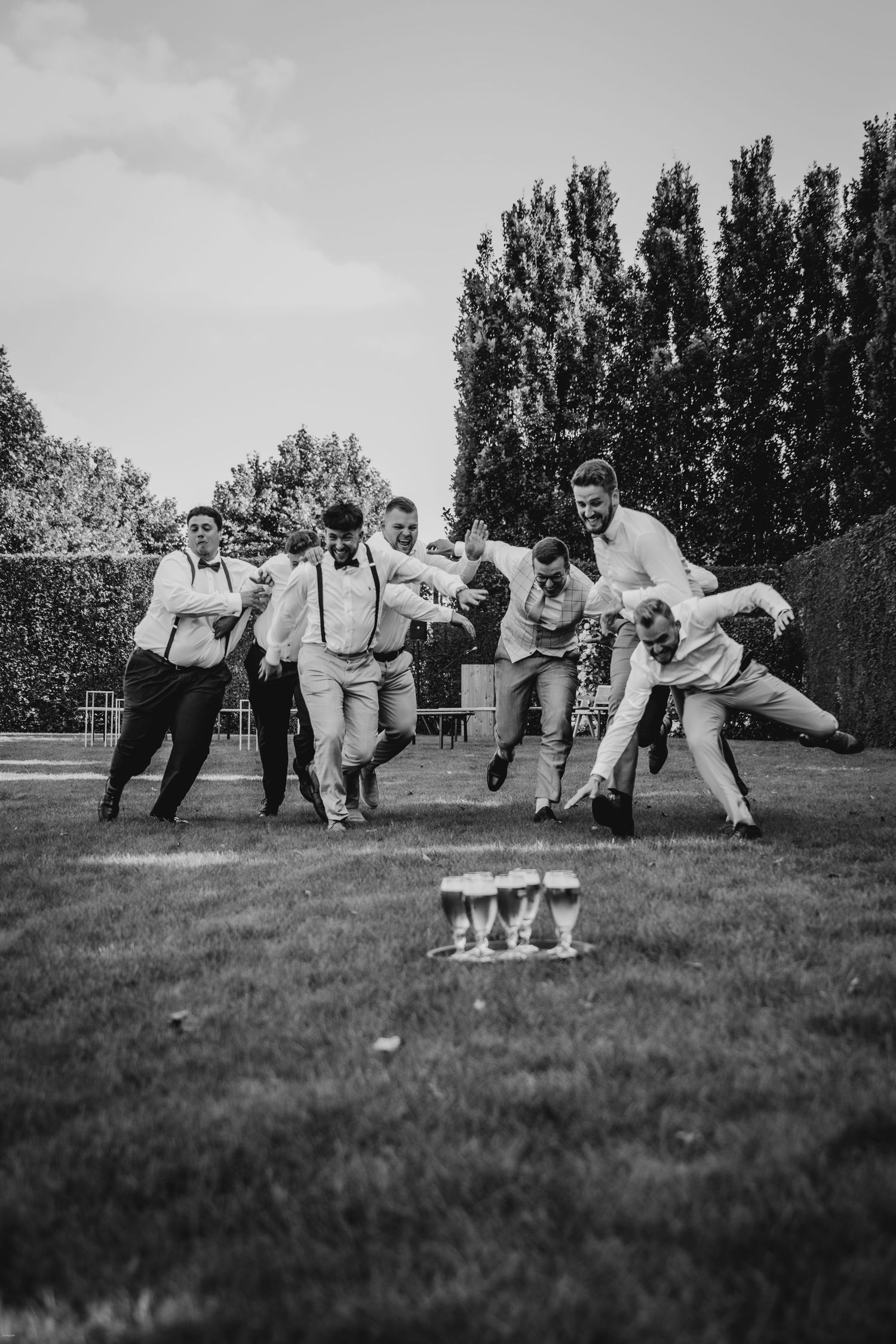 Groomsmen perform acrobatic poses on grass with champagne bottles.