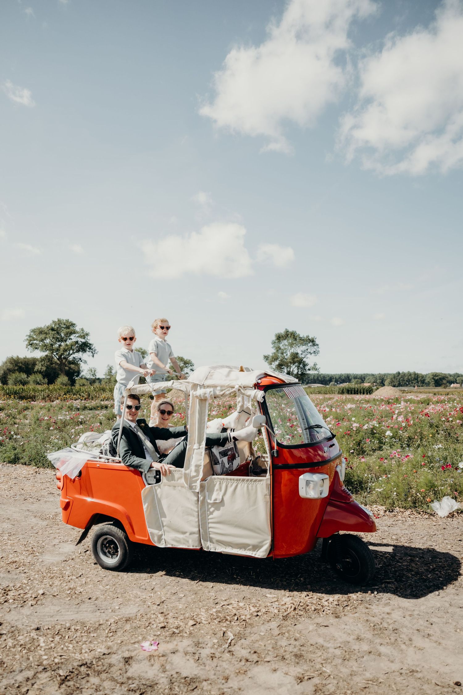 Red and white tuk tuk vehicle carries passengers through a sunny countryside field.