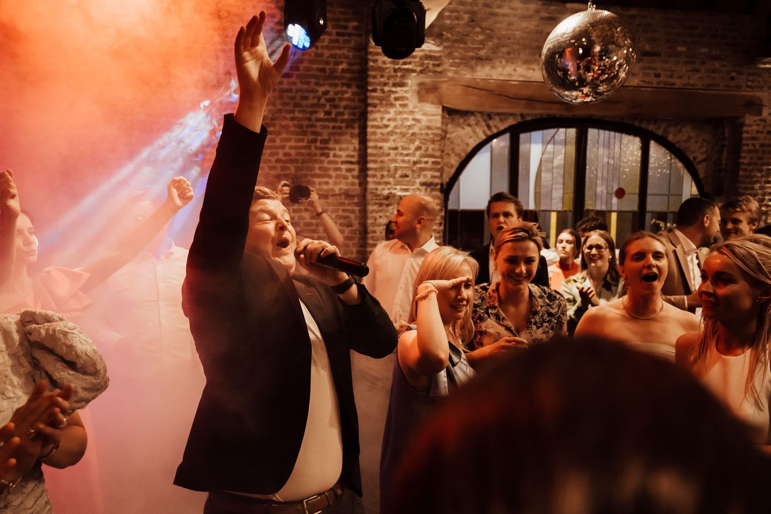 Wedding reception dance party with disco ball and colorful lighting in brick-walled venue.