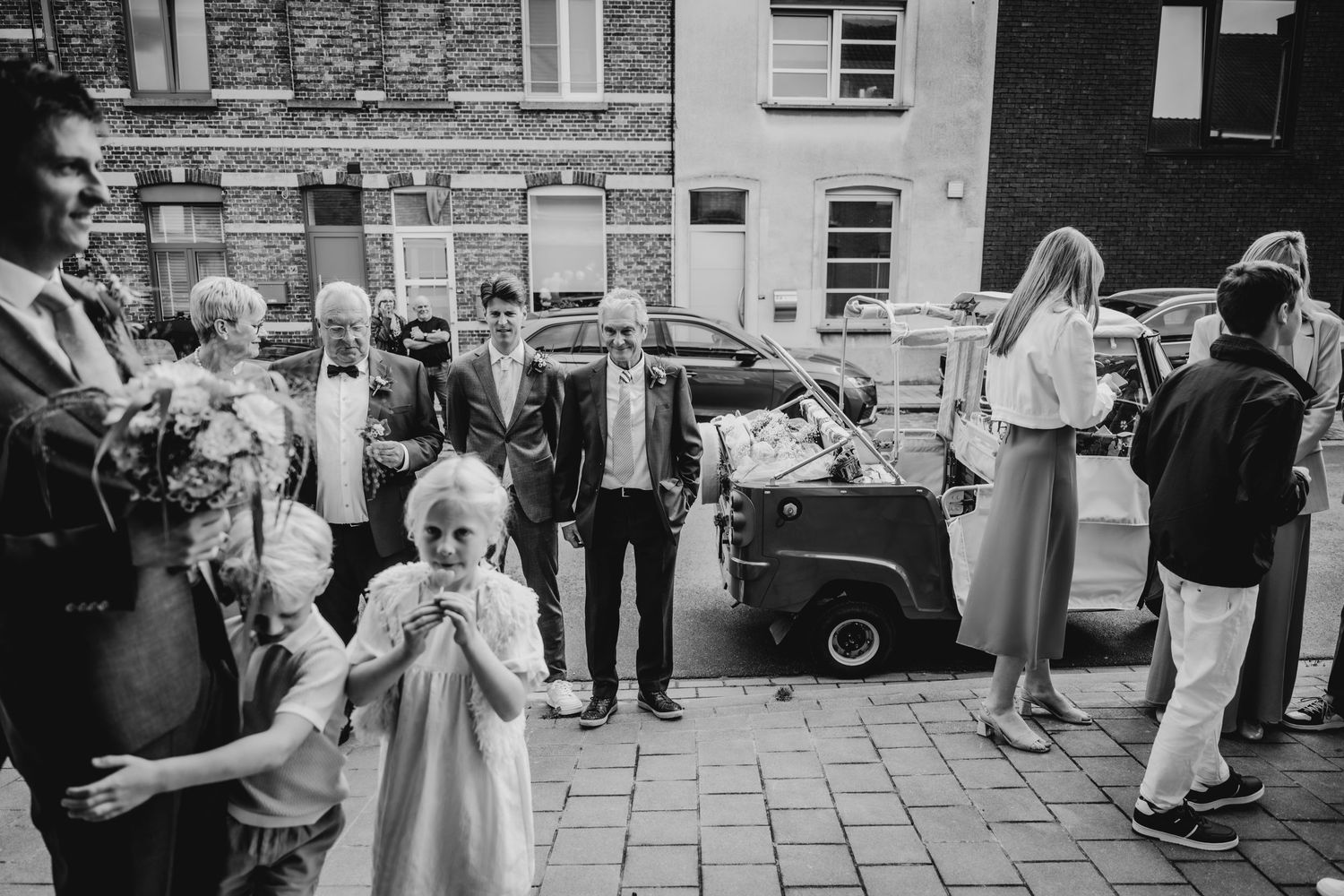 Wedding guests gather outside a brick building on a city street during a celebration.