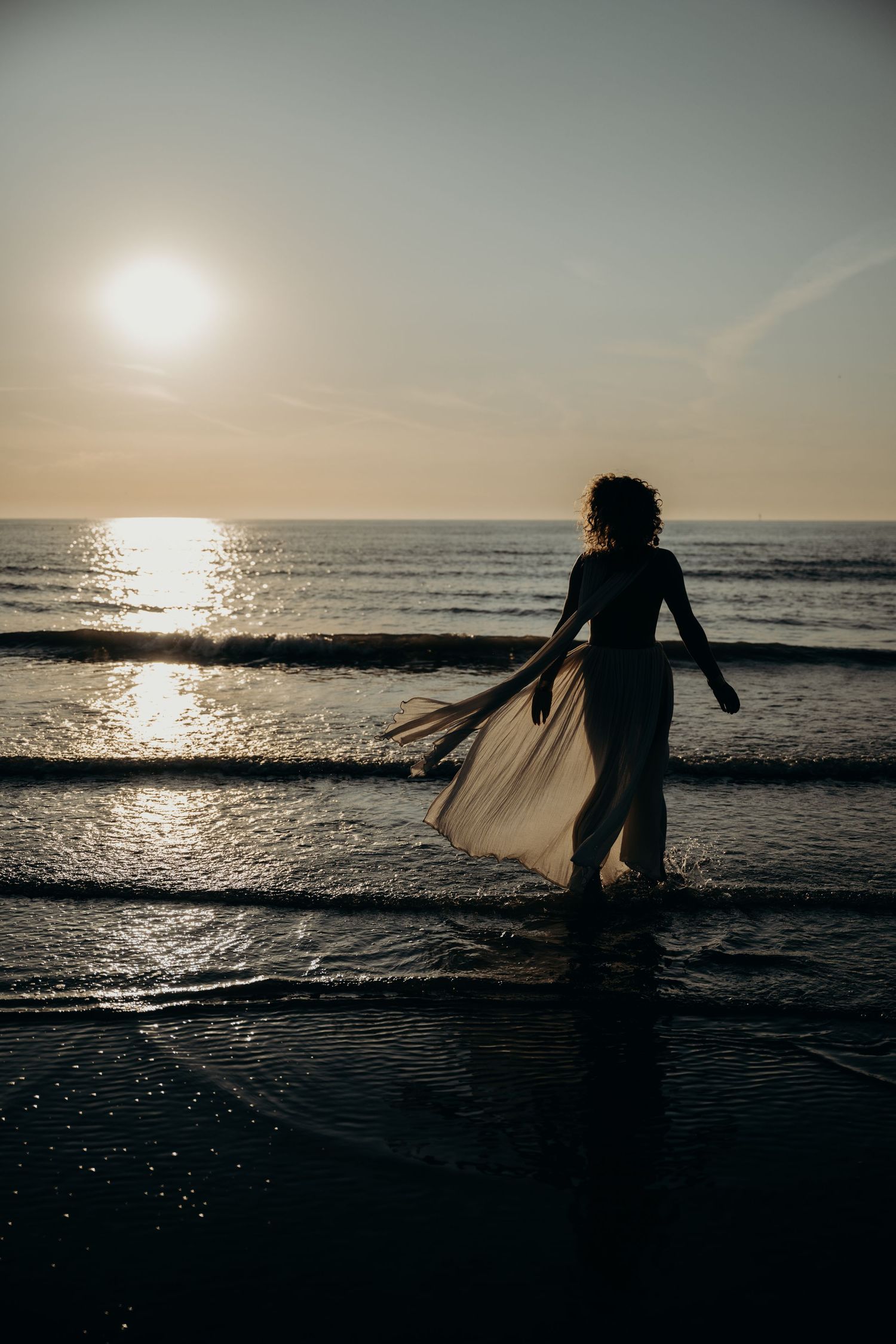 Silhouette of a figure in flowing dress dancing on beach at sunset with ocean waves in background.