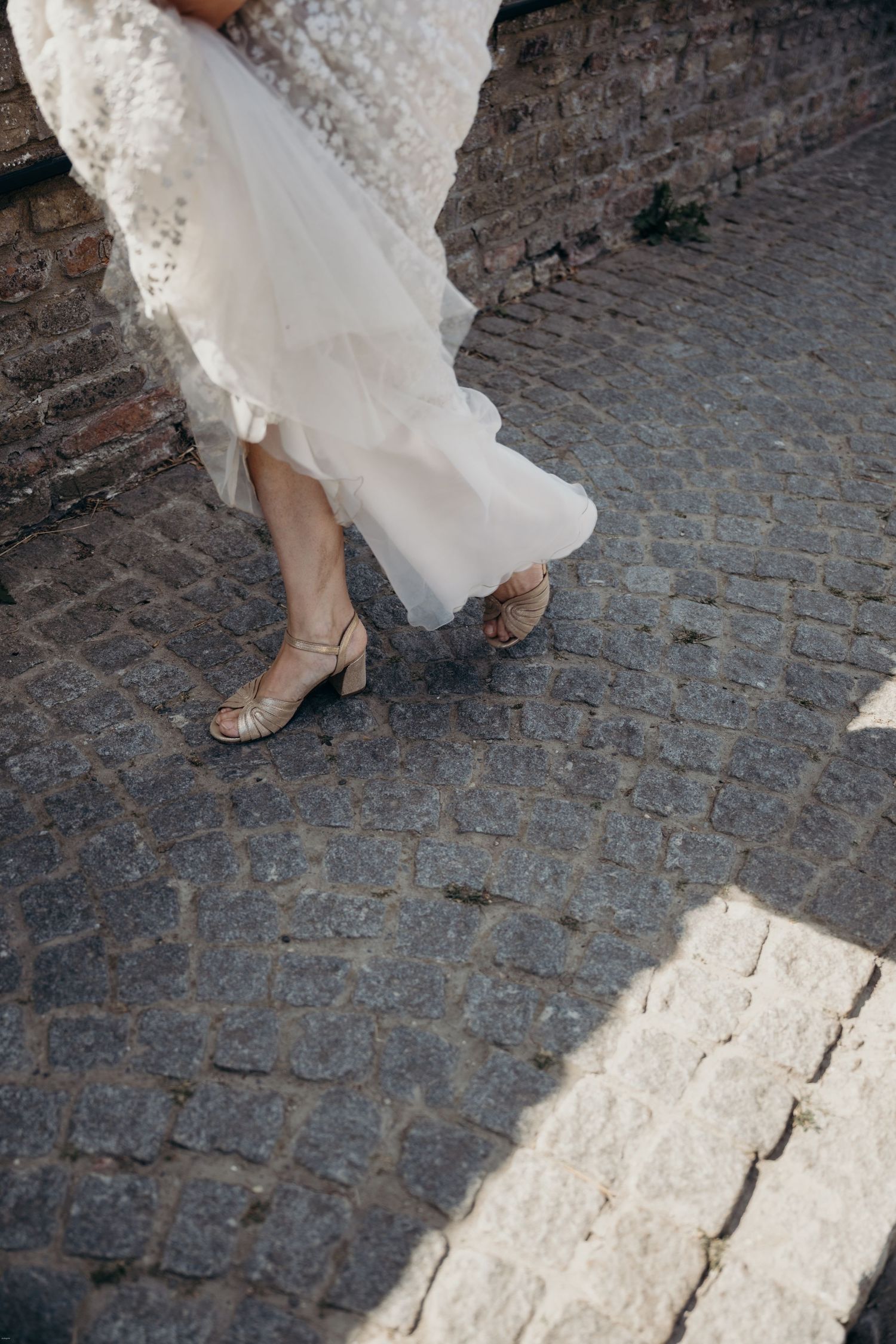 Close-up of elegant bridal shoes and flowing white dress hem on cobblestone pavement.