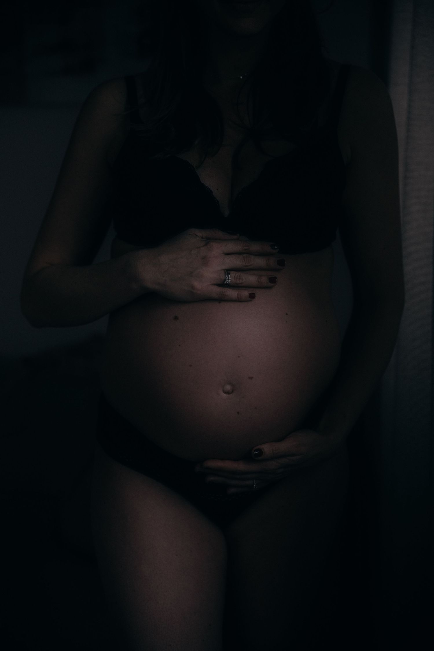A dark and moody maternity photo showing hands cradling a pregnant belly in silhouette against a black background.