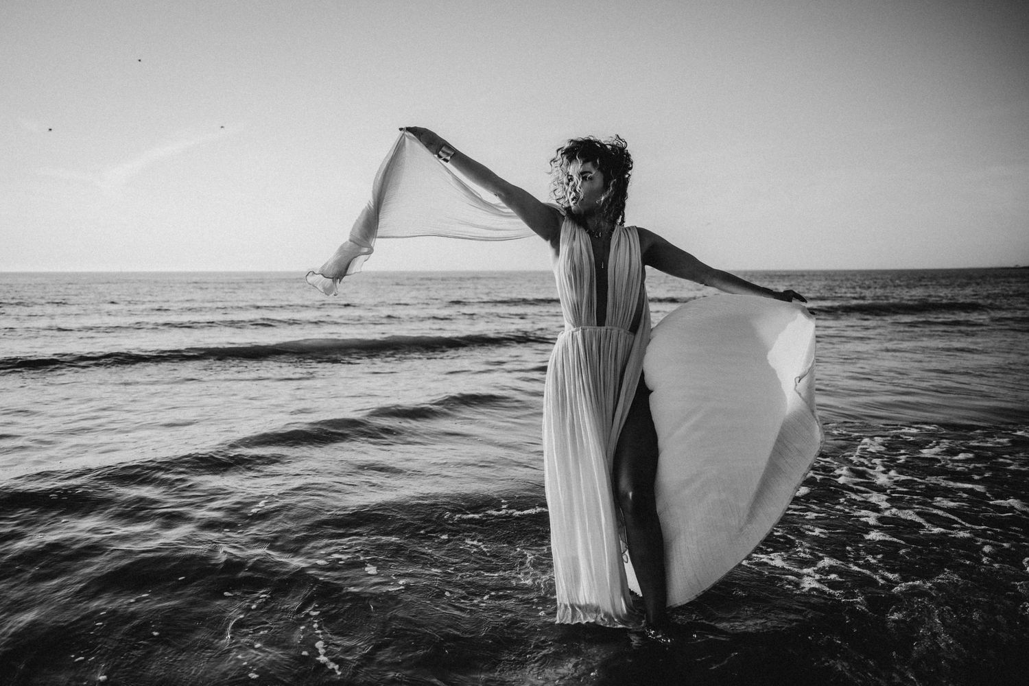 Black and white photo of a figure in flowing white dress dancing on a beach at sunset.