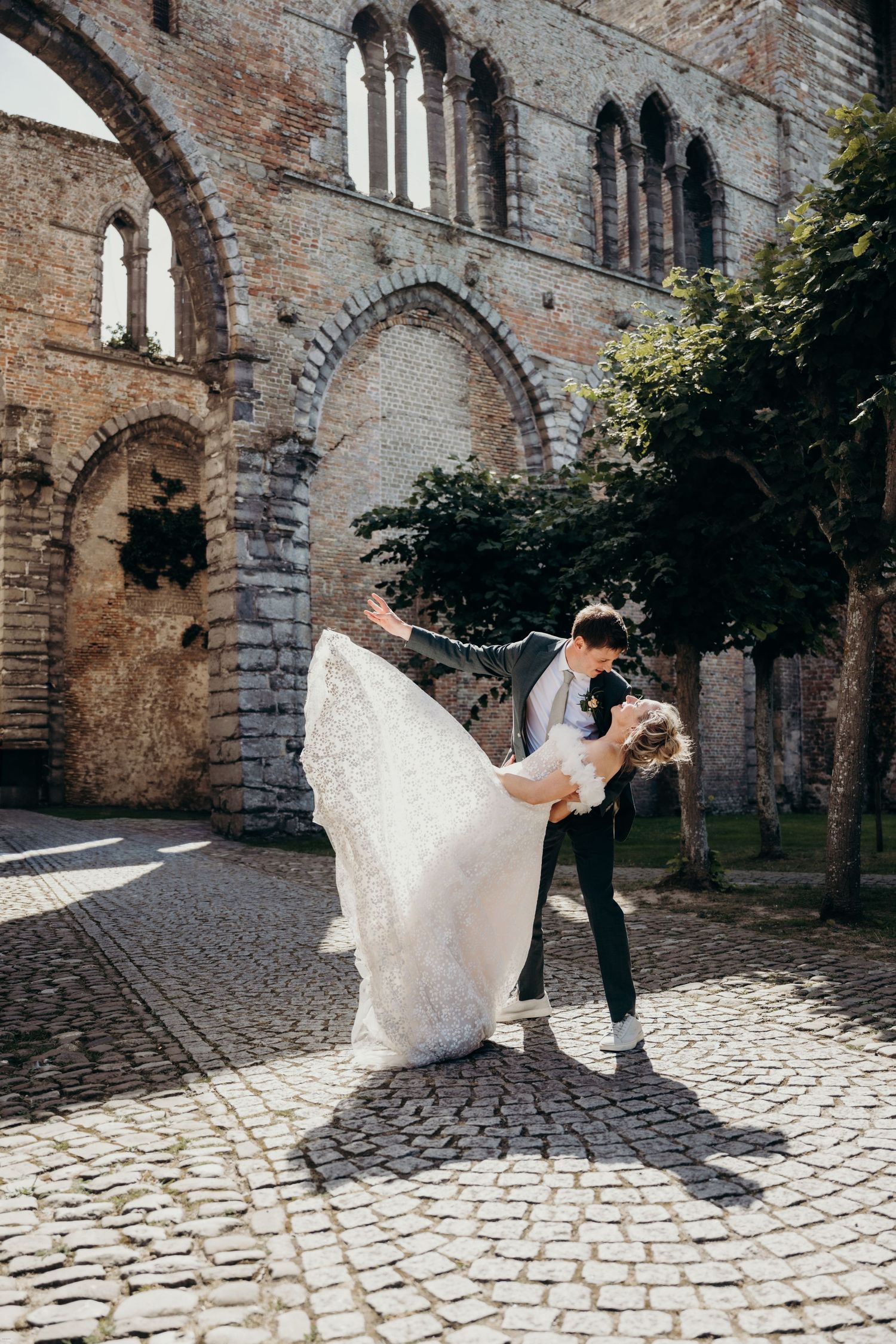 Dancing couple in wedding attire twirls on cobblestone path beside ancient stone church walls.