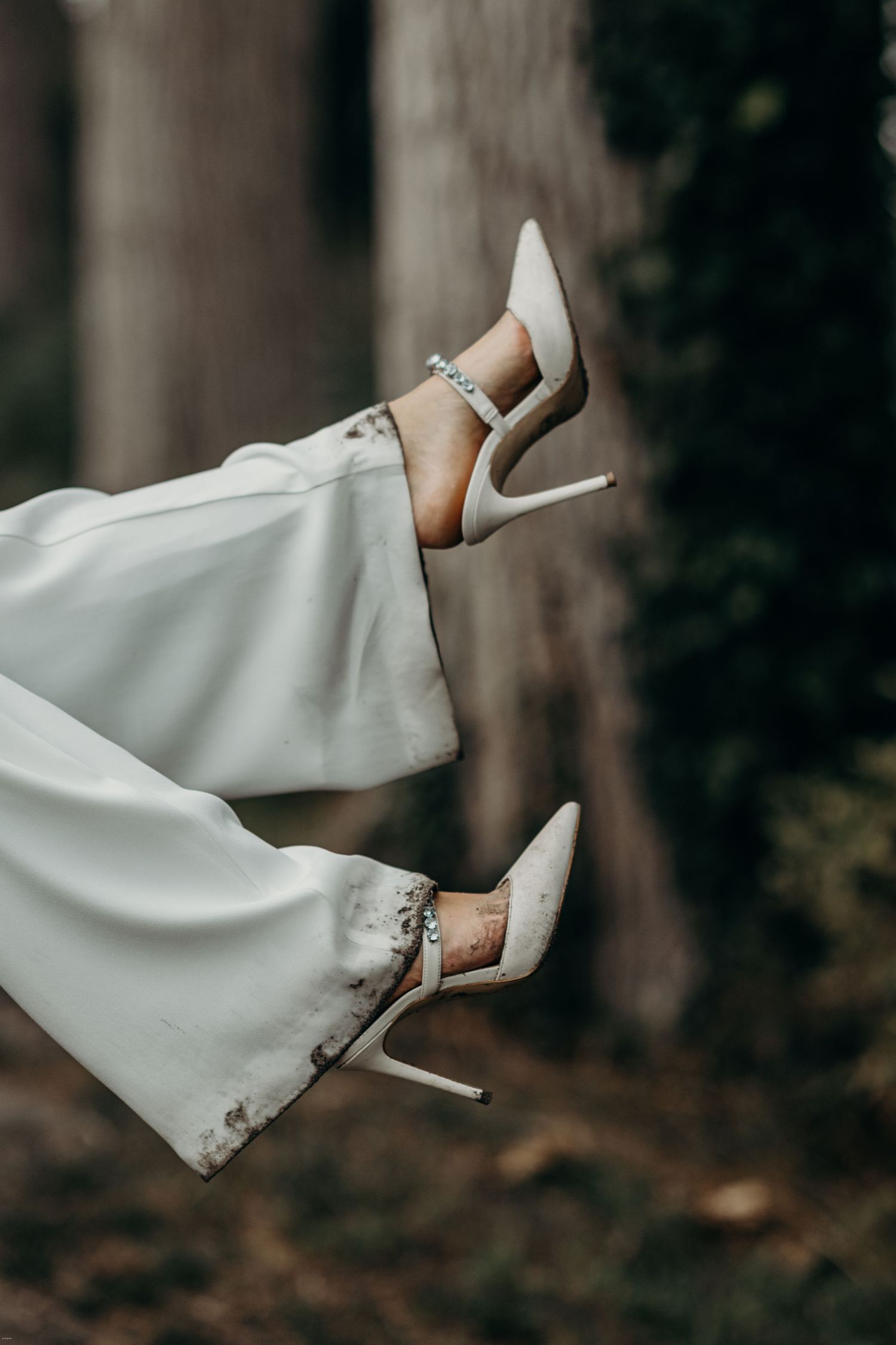 Close-up of white wedding shoes and dress hem in a forest setting.