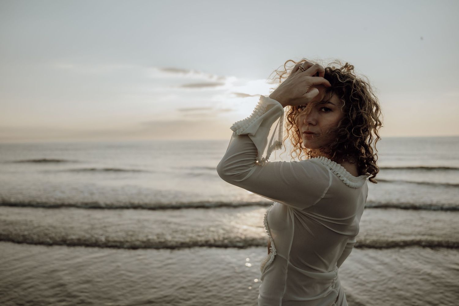 Person in white clothing dancing gracefully on a beach at sunset with ocean waves in the background.