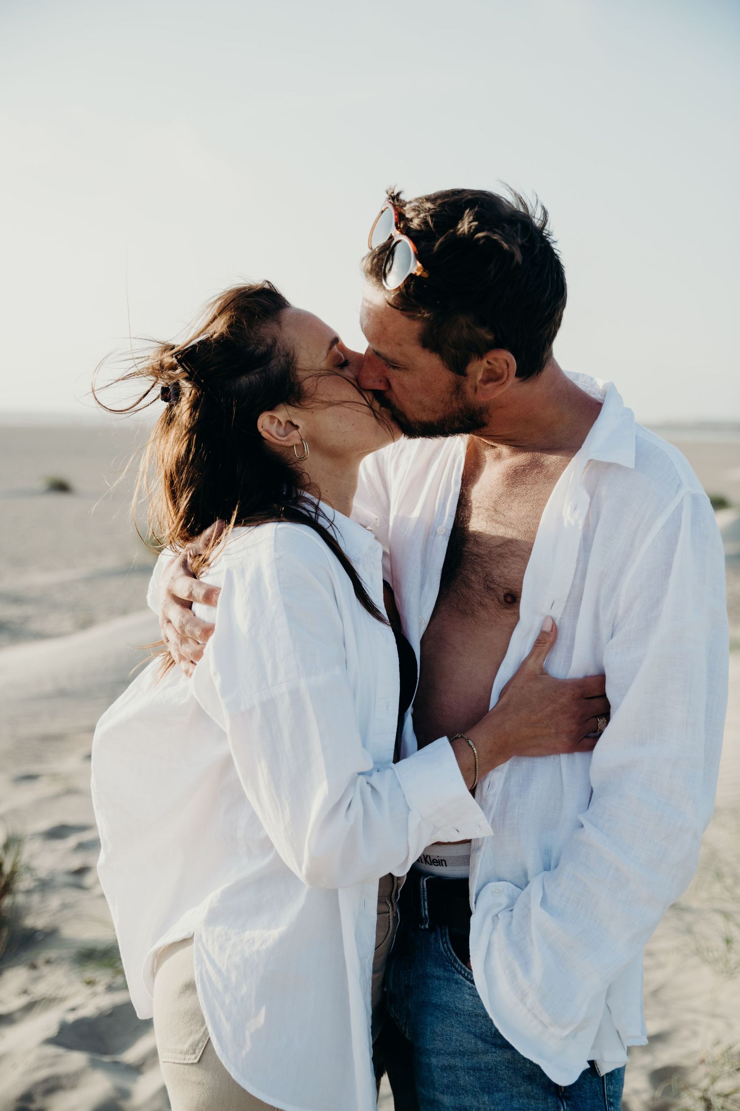 Romantic couple in white shirts share intimate kisses on sandy beach at sunset.