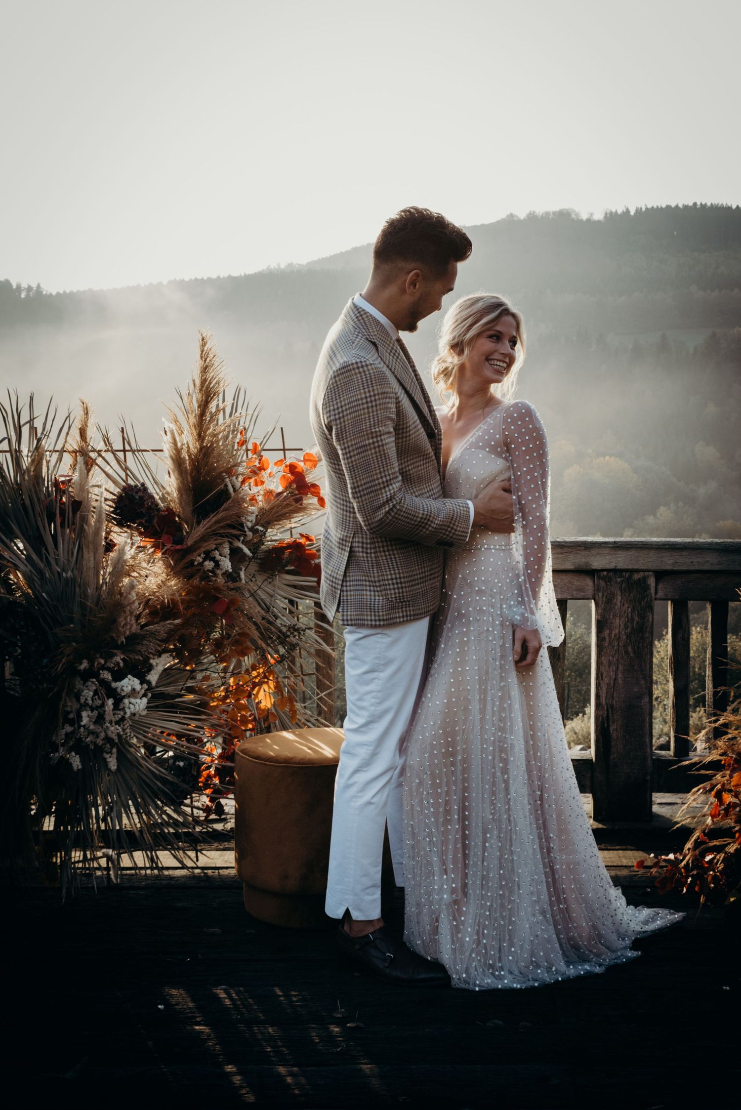 Romantic couple in wedding attire embrace on a rustic deck with mountain views and dried floral arrangements at sunset.