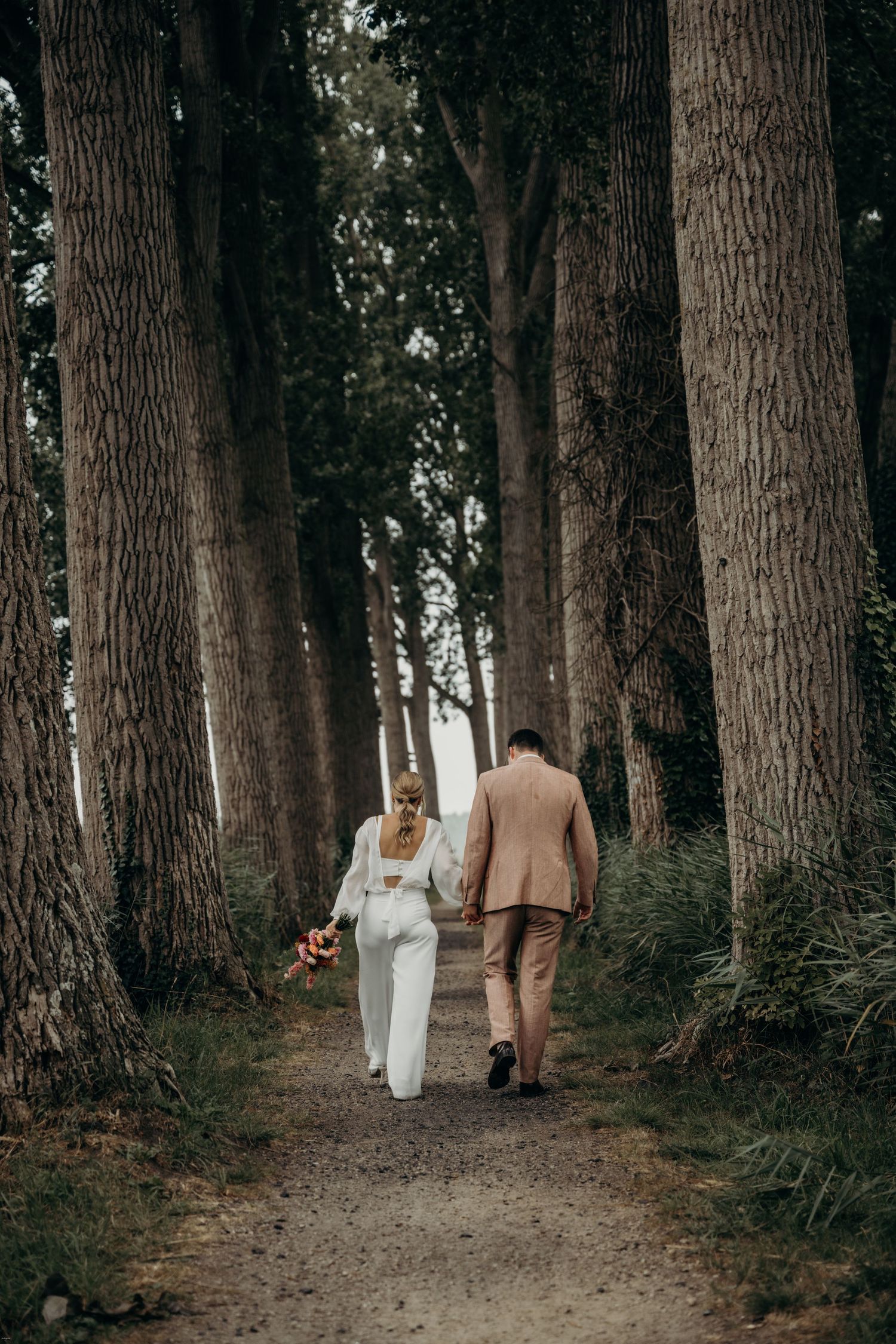 Couple walking hand in hand down a forest path surrounded by tall trees.