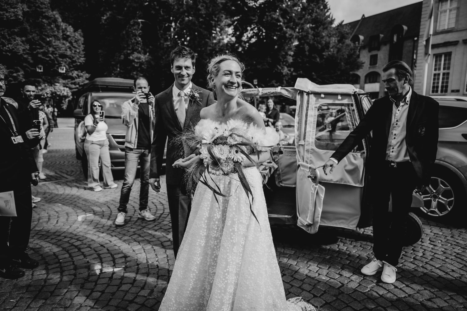 A wedding party walks down a cobblestone street with vintage cars in the background.